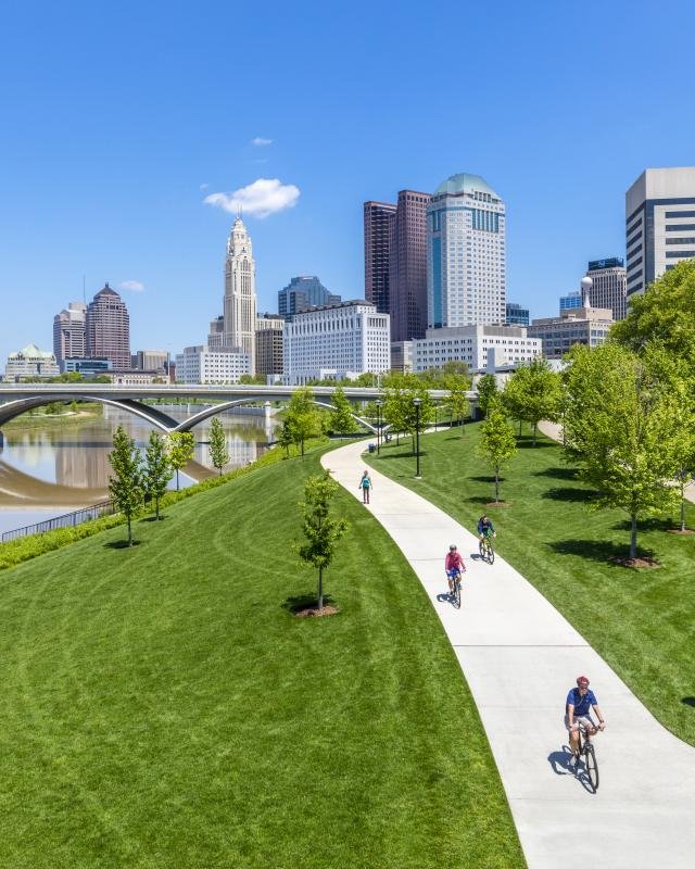 Columbus, OH skyline with tall buildings viewed from a park with a winding pathway, green grass, trees, and people riding bikes on a sunny day.