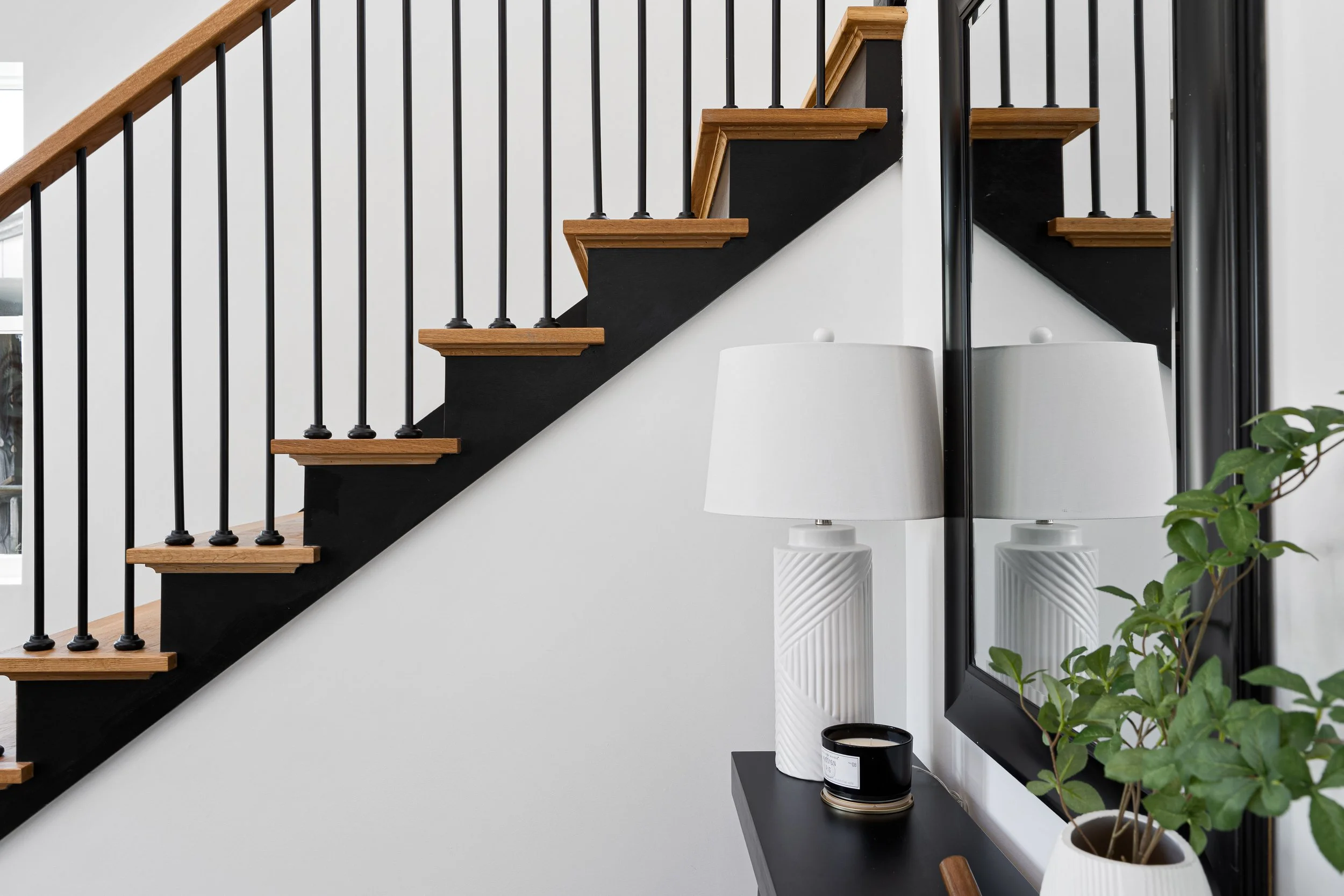 Interior view of a modern staircase with wooden steps and black metal railings, along with a side table with a white table lamp and a potted plant nearby.