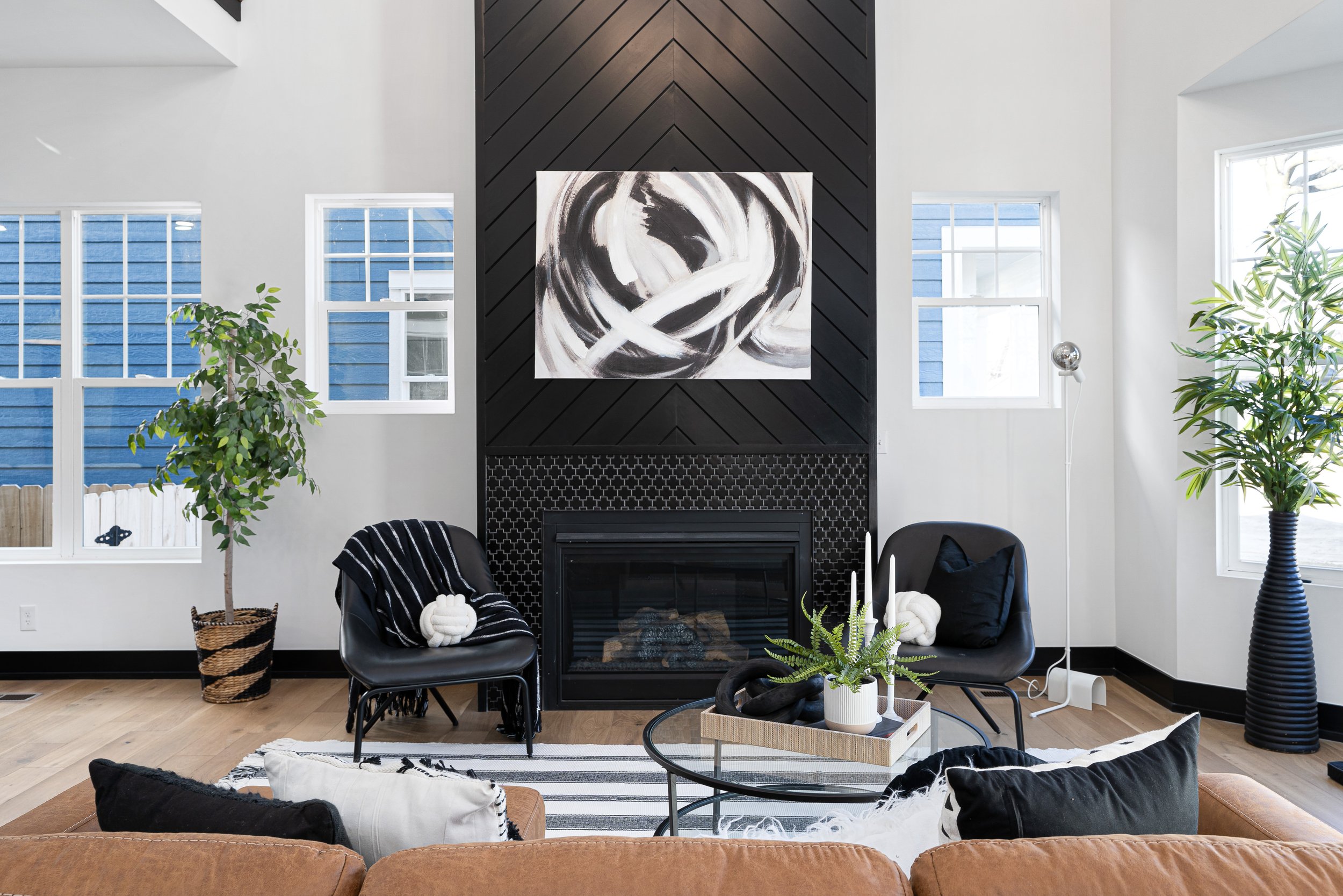 A modern living room with white walls and large windows, a black fireplace with a white and black abstract painting above it, two black chairs with white pillows, a brown sofa, and various green plants in baskets and pots.