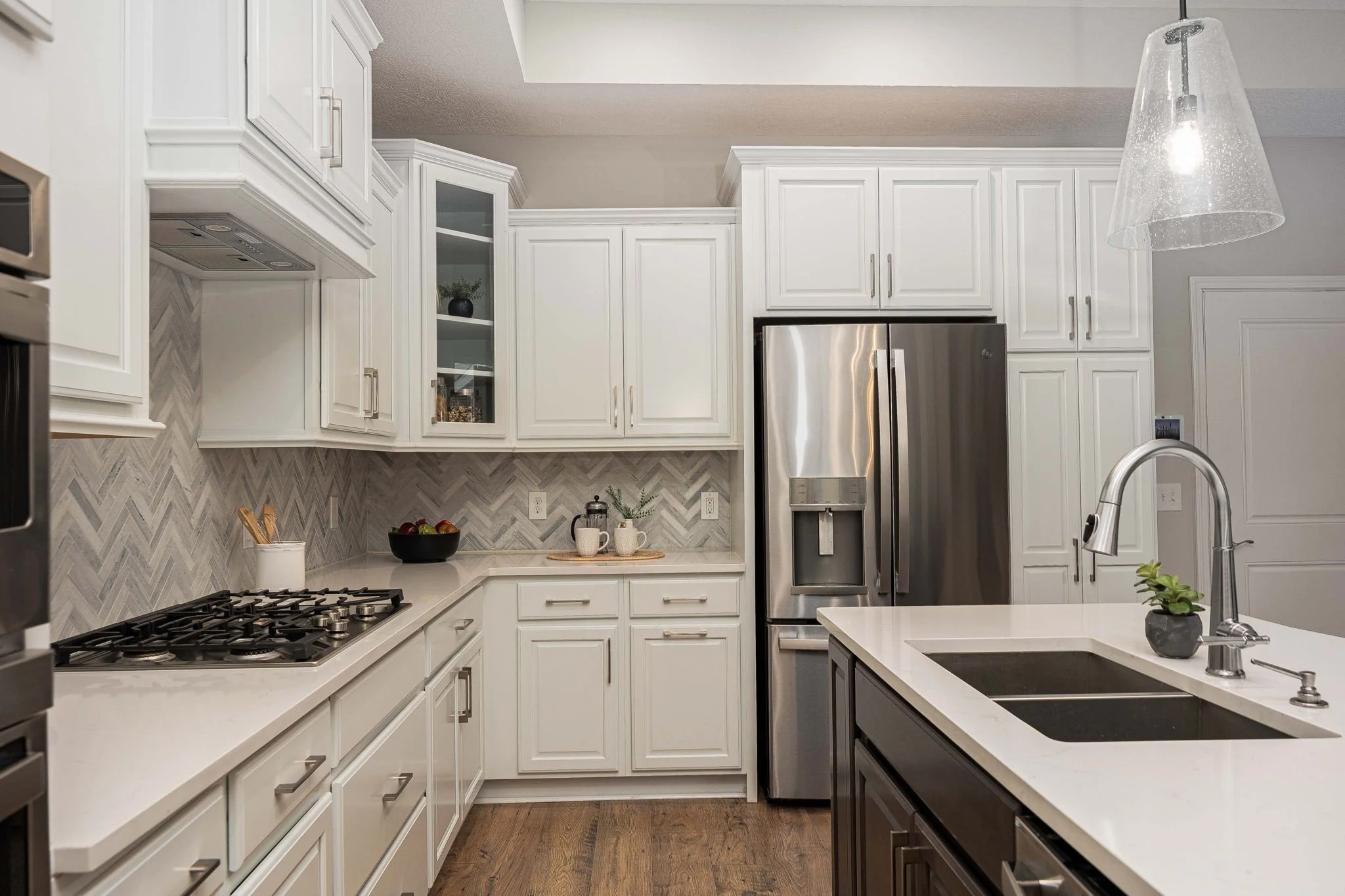 Modern kitchen with white and dark cabinetry, stainless steel refrigerator, countertop with fruit and coffee cups, and a pendant light fixture.