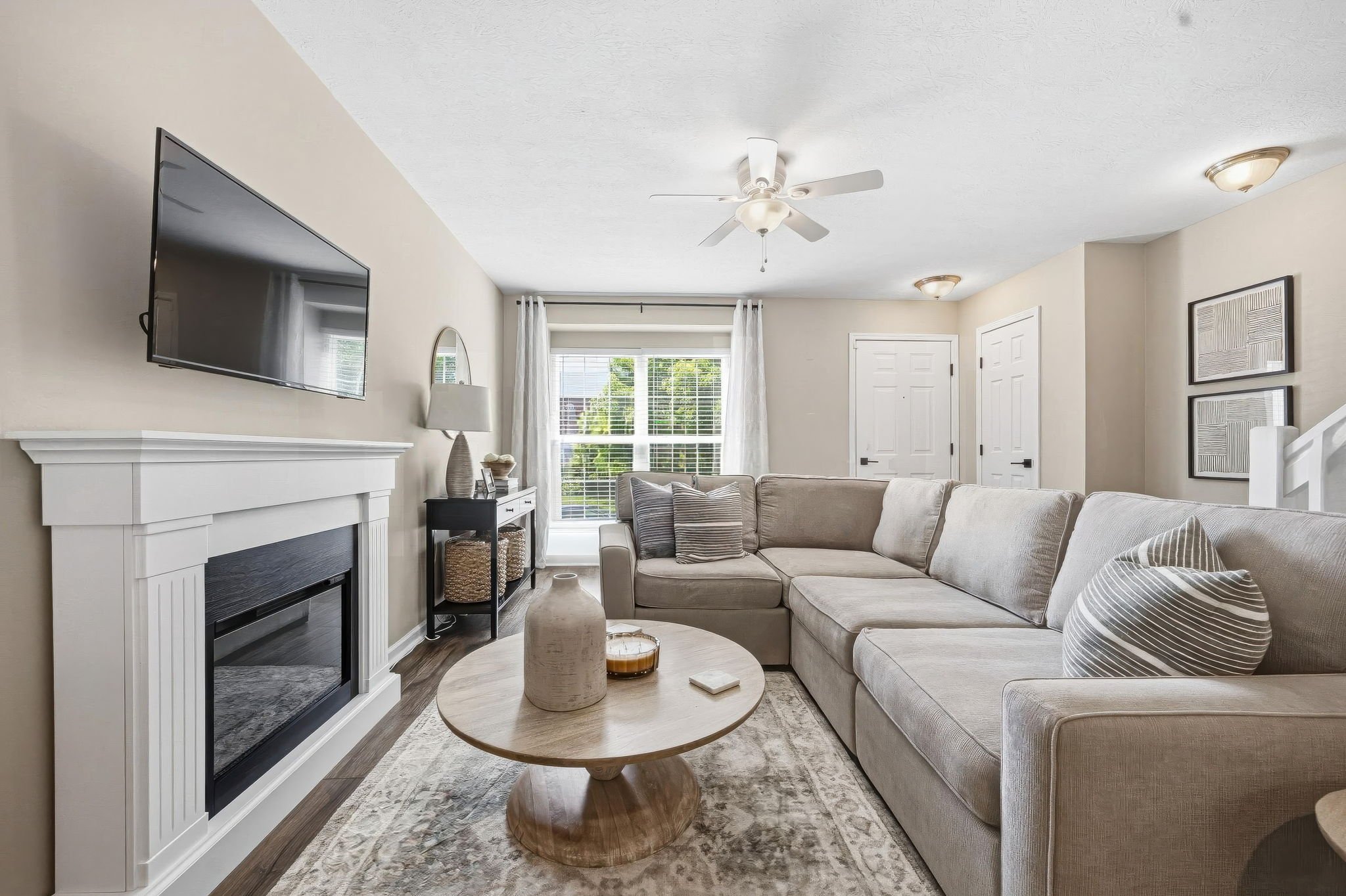 A living room with a beige sectional sofa, two patterned throw pillows, a round wooden coffee table, a fireplace with a white mantel, a wall-mounted flat-screen TV, a coffee table with decorative vases, a window with white blinds and curtains, framed artwork on the wall, a ceiling fan, and ceiling lights.