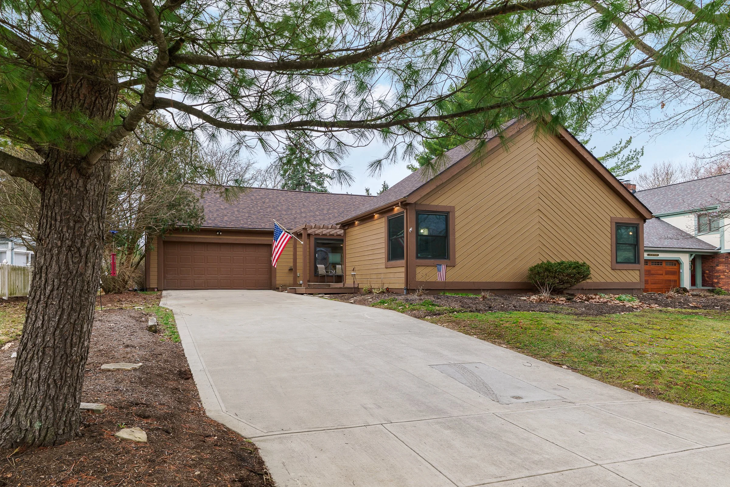 A yellow house with a brown garage door, two windows, and a small porch, situated under a large leafy tree with a driveway leading up to it, with grass and shrubs around.