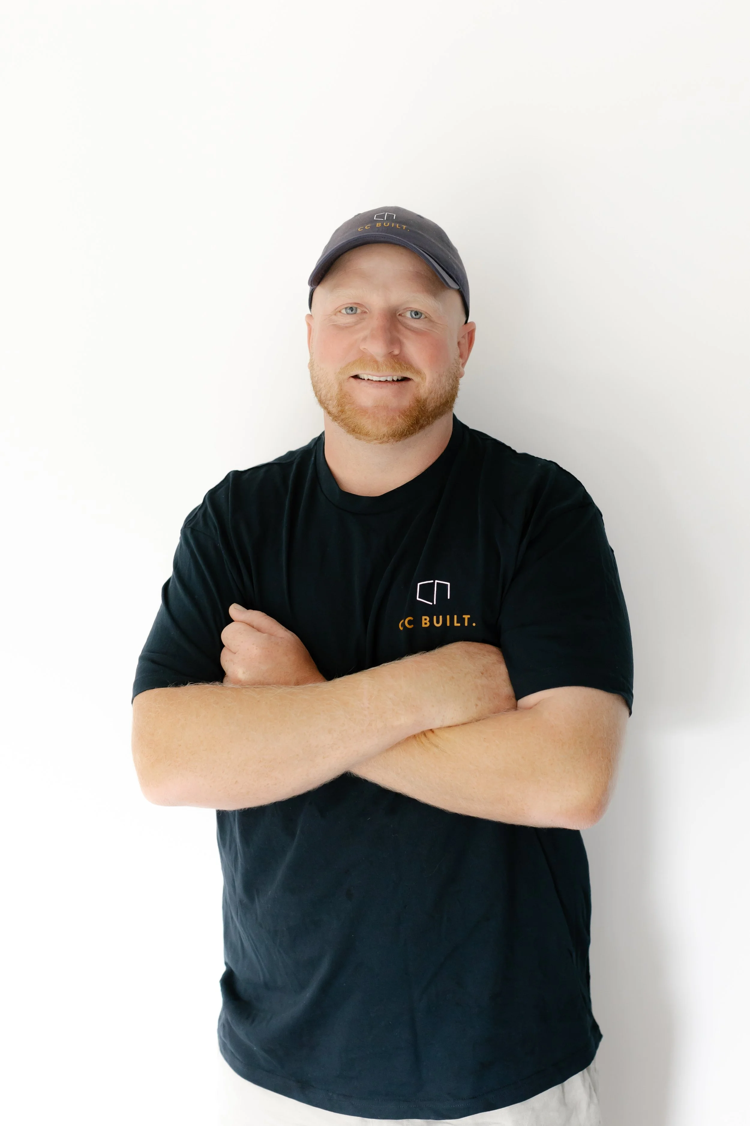 A man with a ginger beard and blue eyes is smiling, wearing a black T-shirt and a dark baseball cap, standing with arms crossed against a white background.
