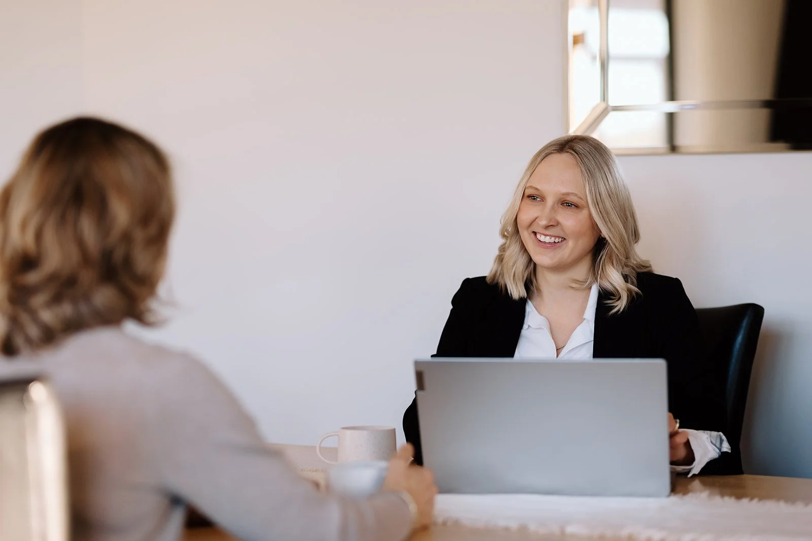 Two women having a meeting in an office, smiling at each other. One woman is in the foreground with blurred hair, and the other woman is in focus, sitting behind a laptop.