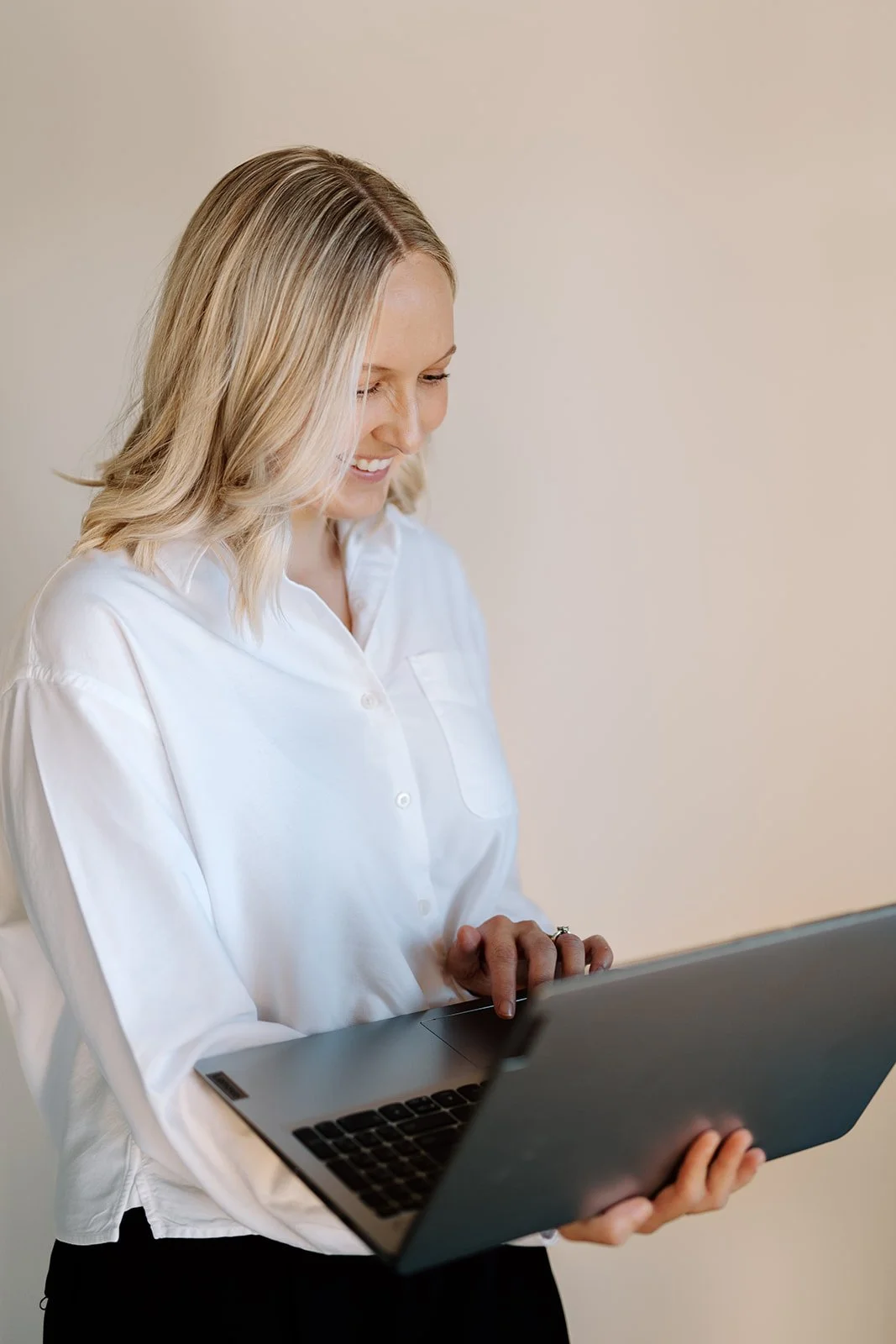 A woman with blonde hair wearing a white shirt, smiling, and looking down at a laptop.