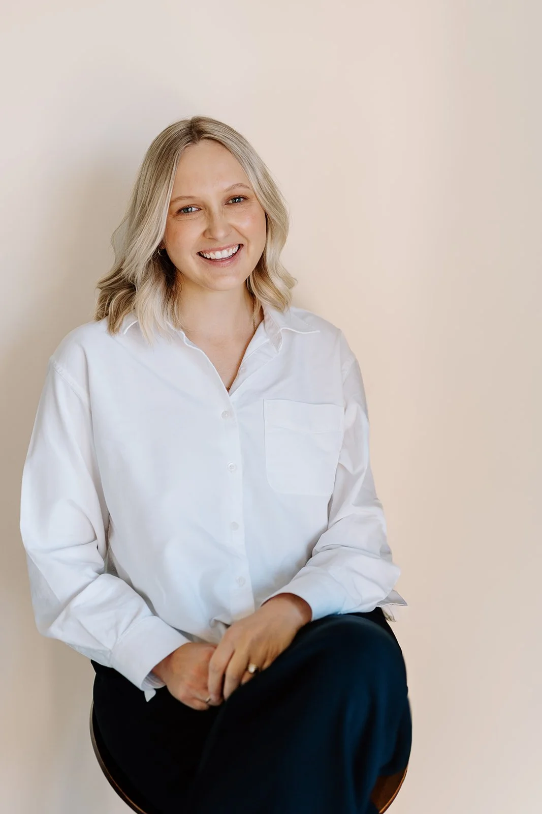 A woman with blonde hair wearing a white shirt sitting against a plain beige wall, smiling at the camera.