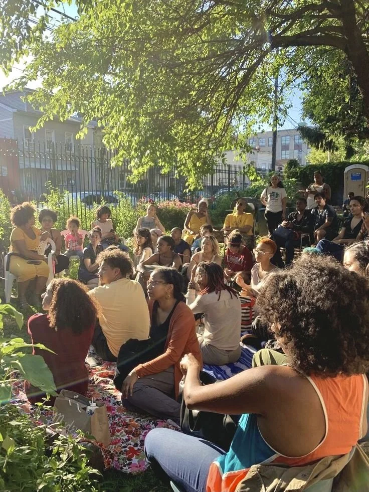 A group of diverse people gathered outdoors in a garden with green trees and plants, sitting on chairs and blankets, listening to someone talk during a sunny spring or summer day.