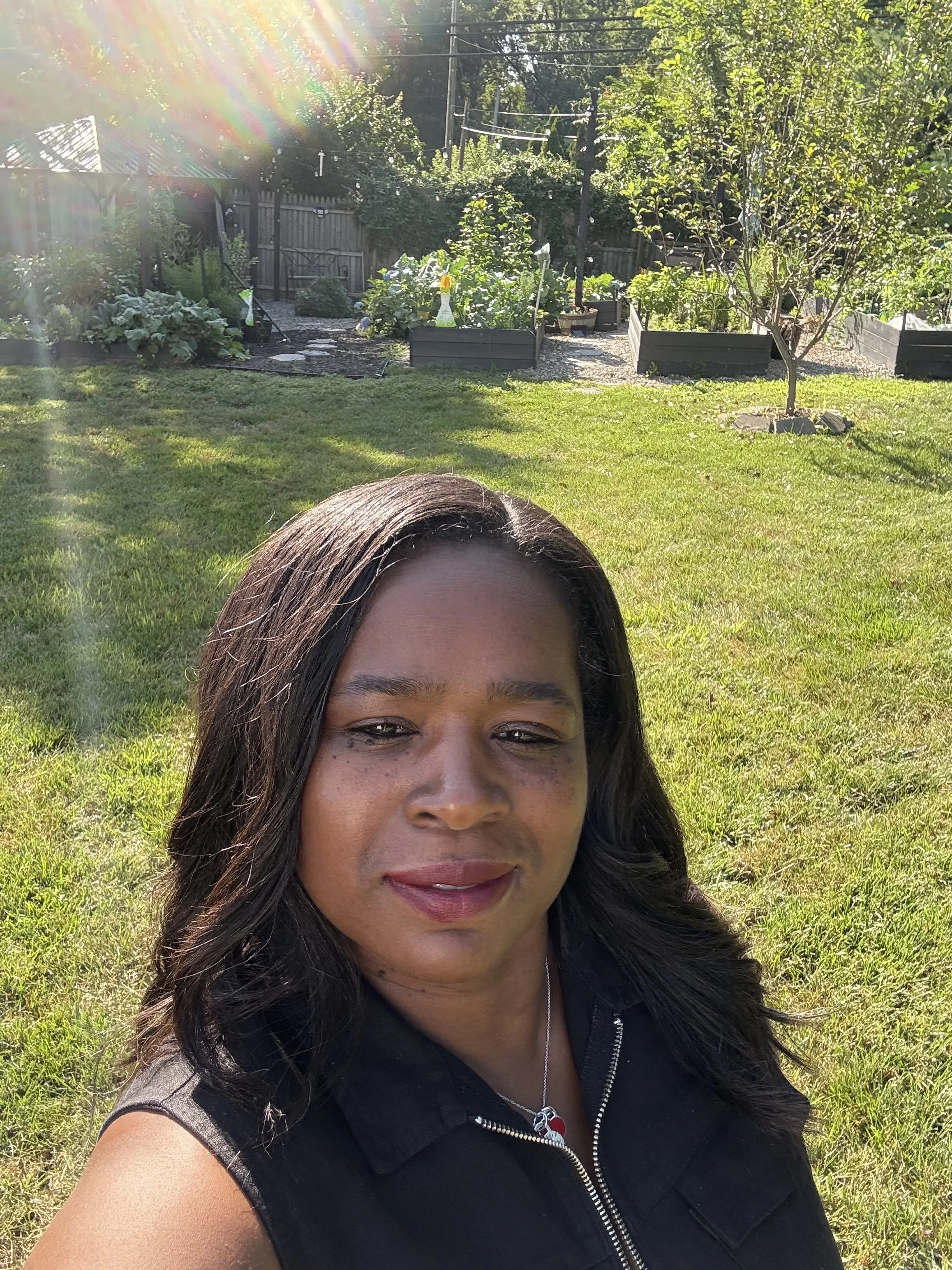 A woman with long dark hair taking a selfie in a sunny garden with green grass, trees, and raised garden beds in the background.