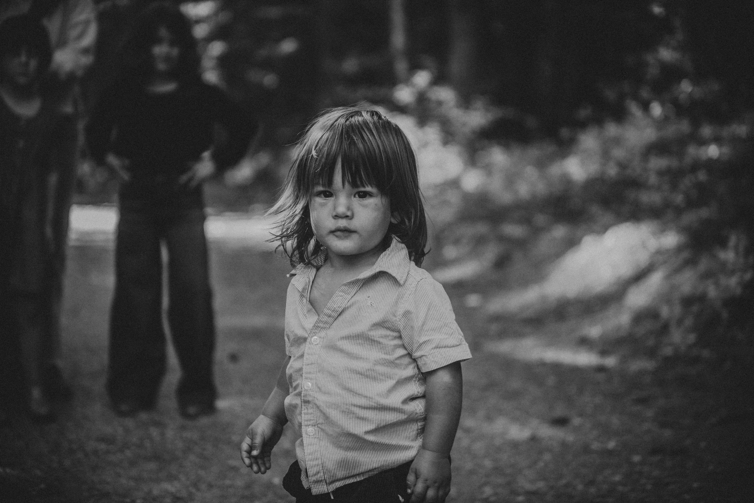 Black and white photo of a young girl with shoulder-length hair standing outdoors, with a neutral expression. Three people are blurred in the background.