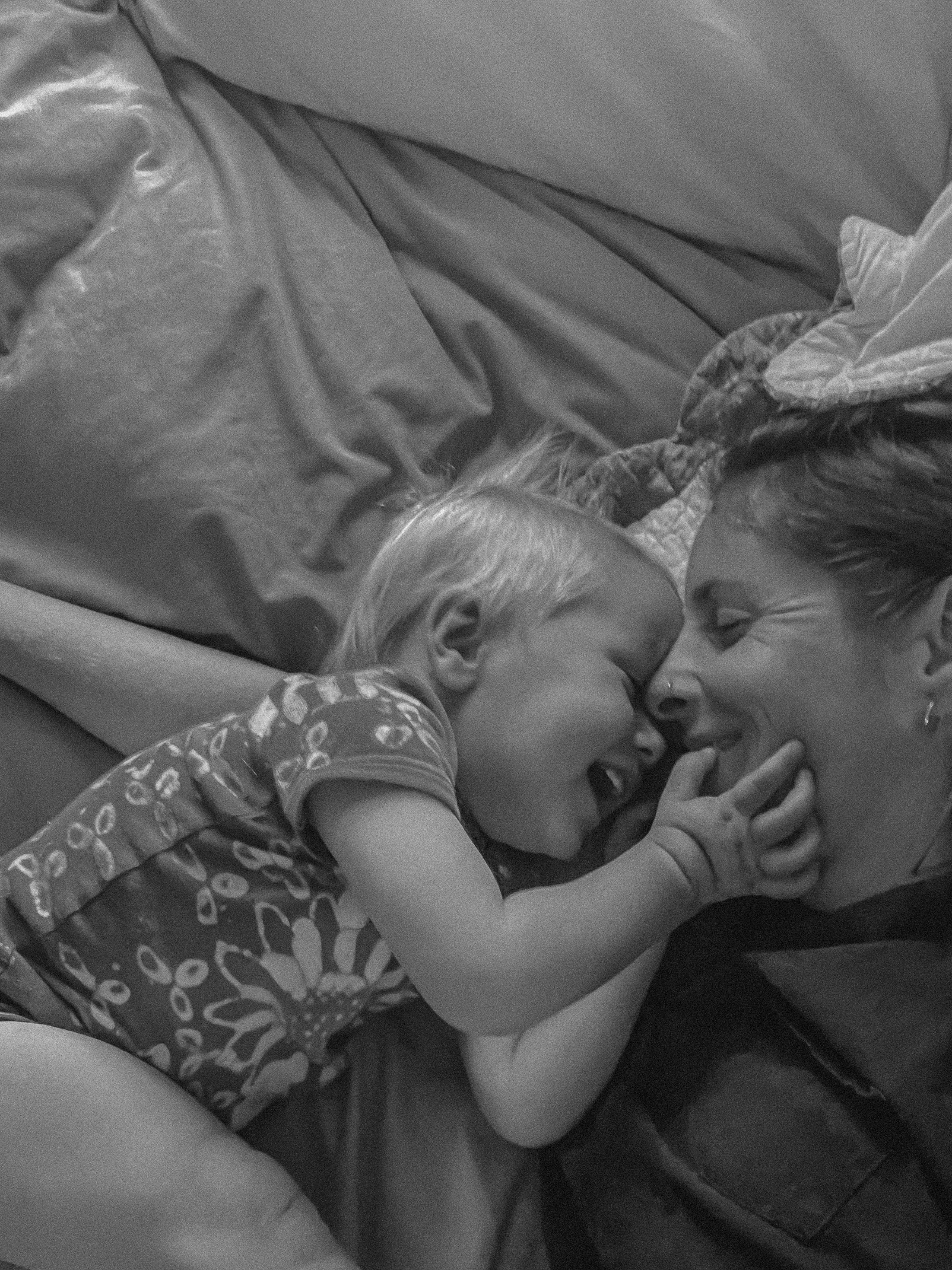 A young girl lying in a bed, snuggling with a woman, presumably her mother. Both are sleeping peacefully with their faces close and eyes closed.
