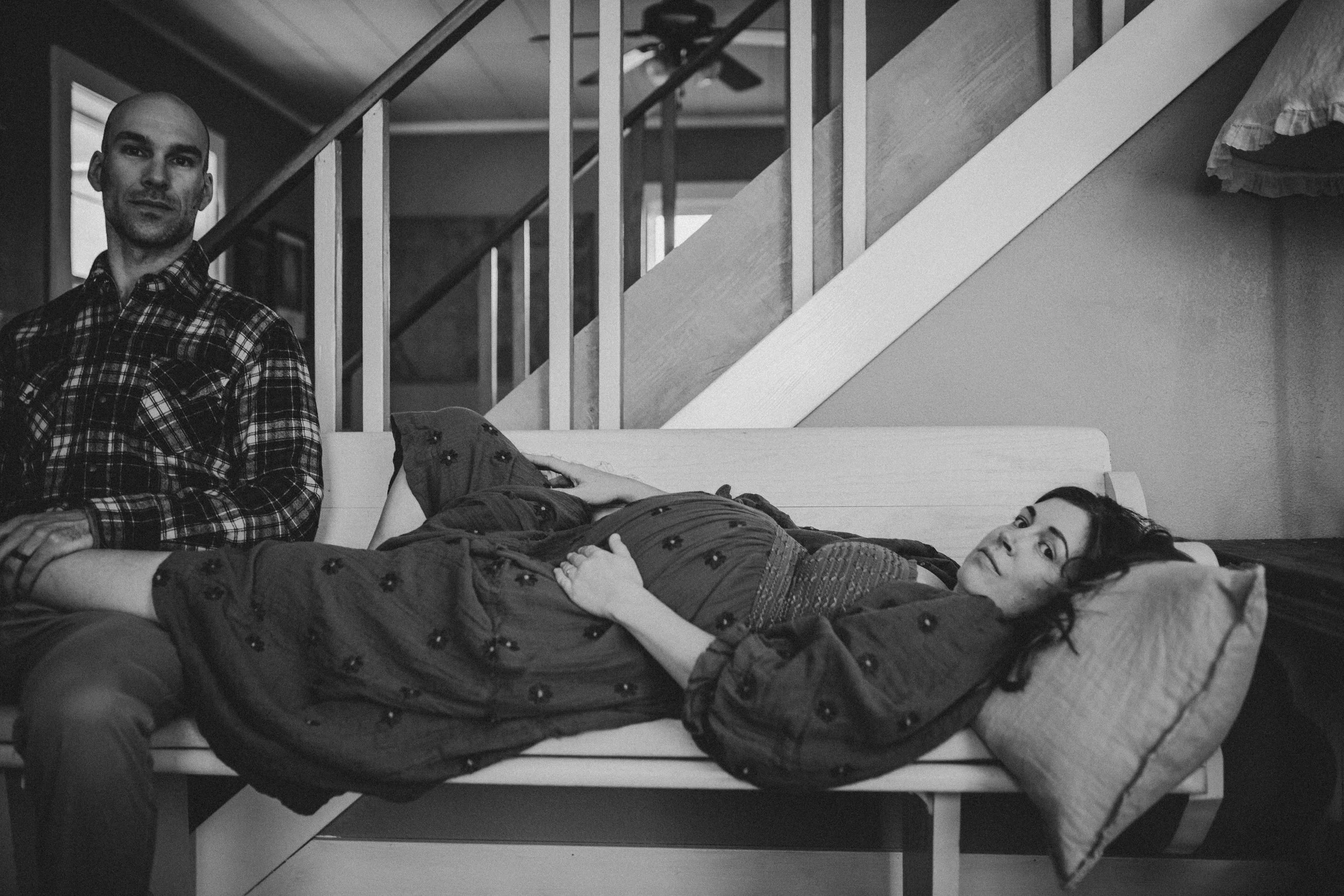 A black-and-white photo of a man with a shaved head and checkered shirt sitting on a bench next to a young woman lying on the same bench. The woman is lying on her side with her head resting on her hand, looking at the camera, smiling. The background