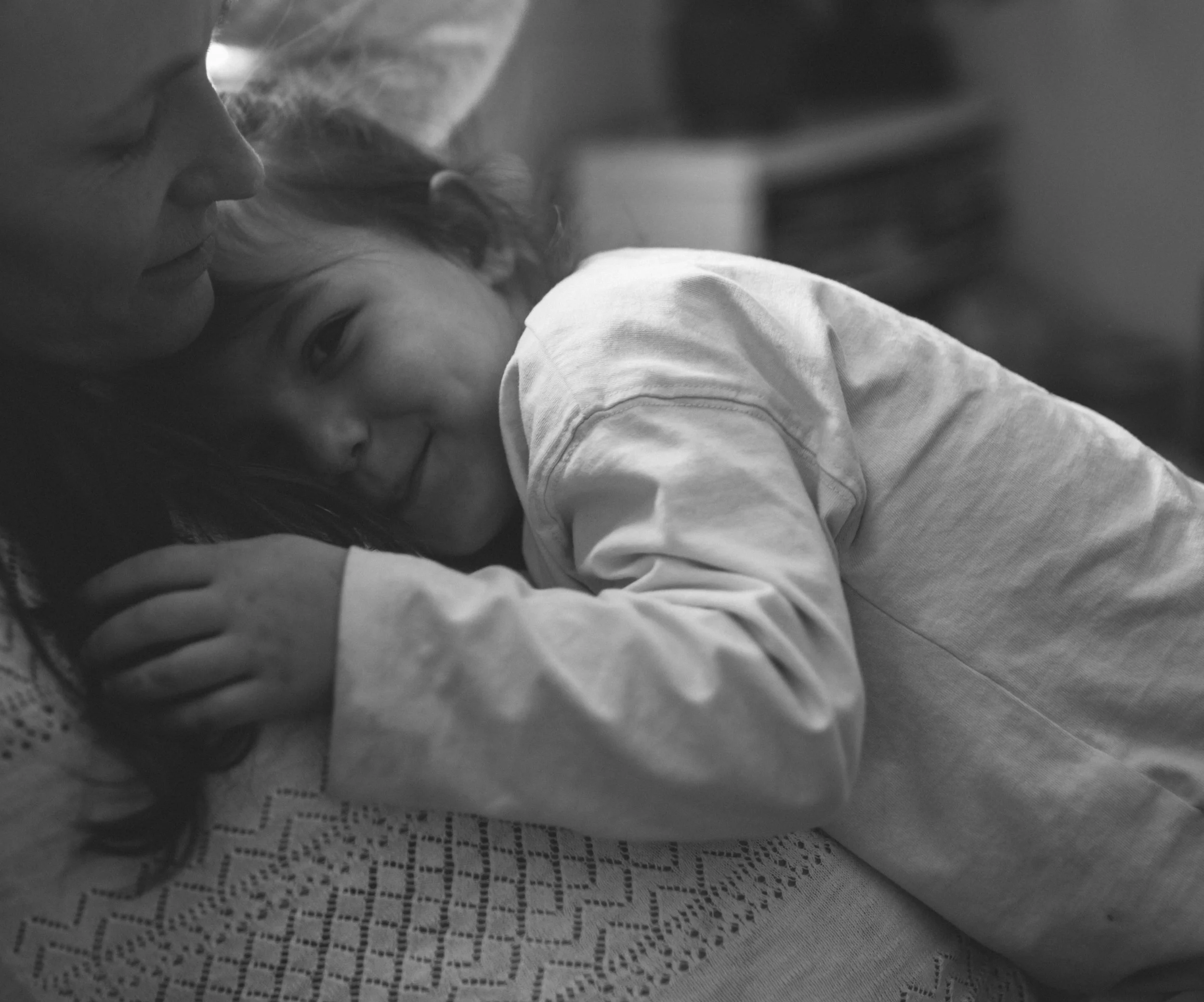 Close-up black and white photo of a woman resting her head on a young child's shoulder, both with closed eyes, tender expression, and lying on a surface with lace fabric.