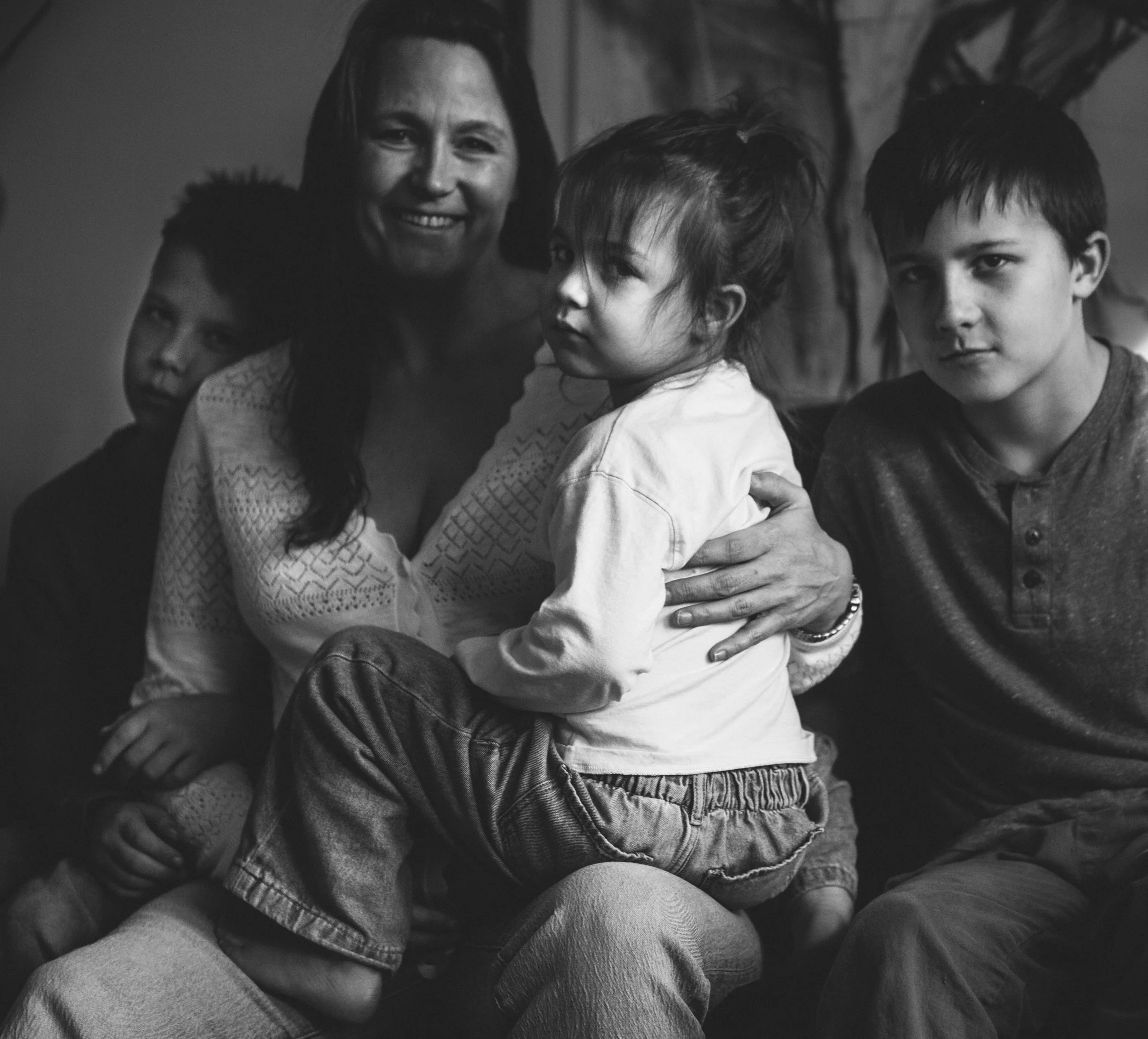 A black and white photo of a group of children sitting closely together, with an adult woman smiling in the background.