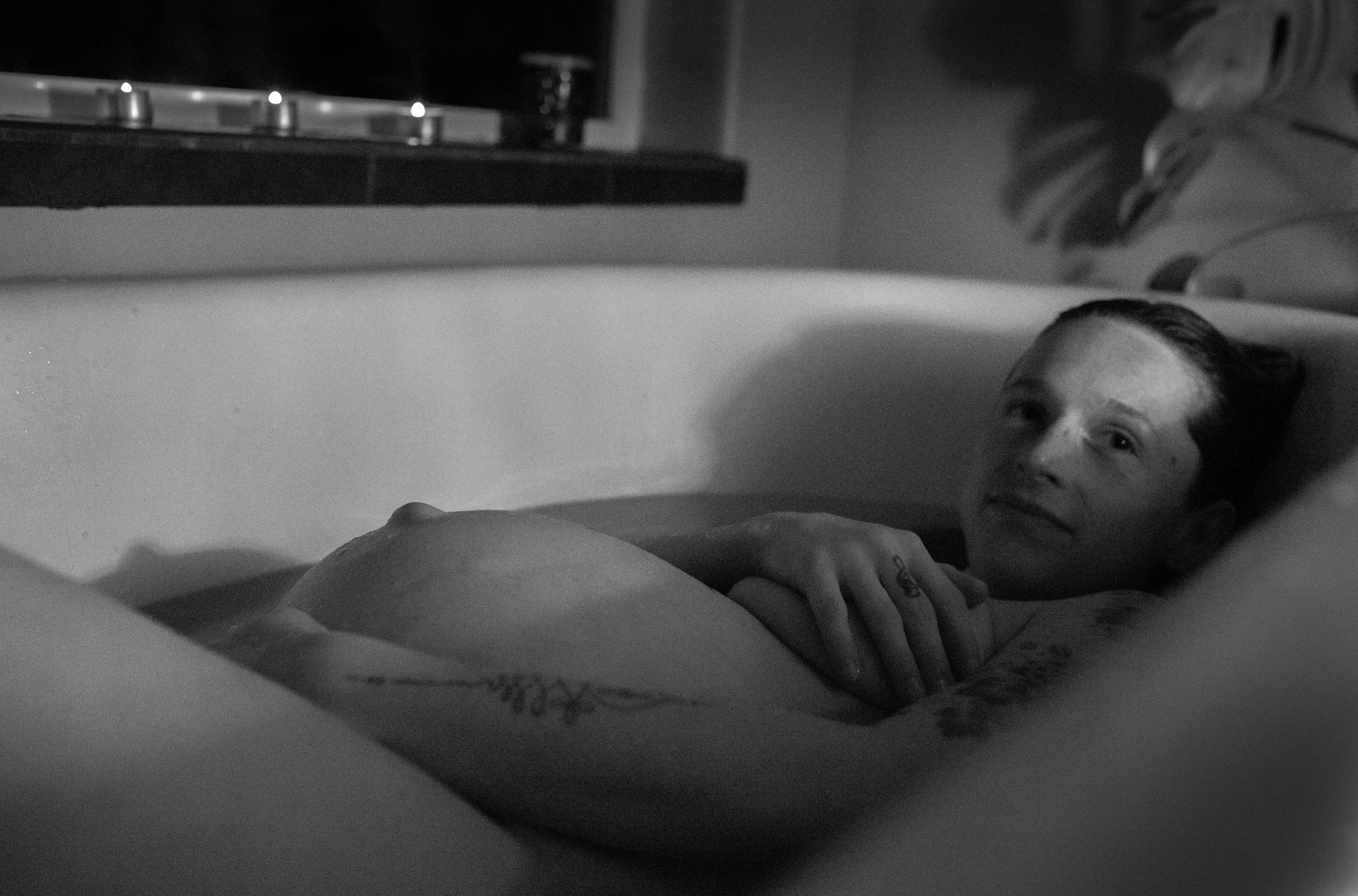 A black-and-white photo of a woman with dark hair relaxing in a bathtub, looking up with a calm expression.