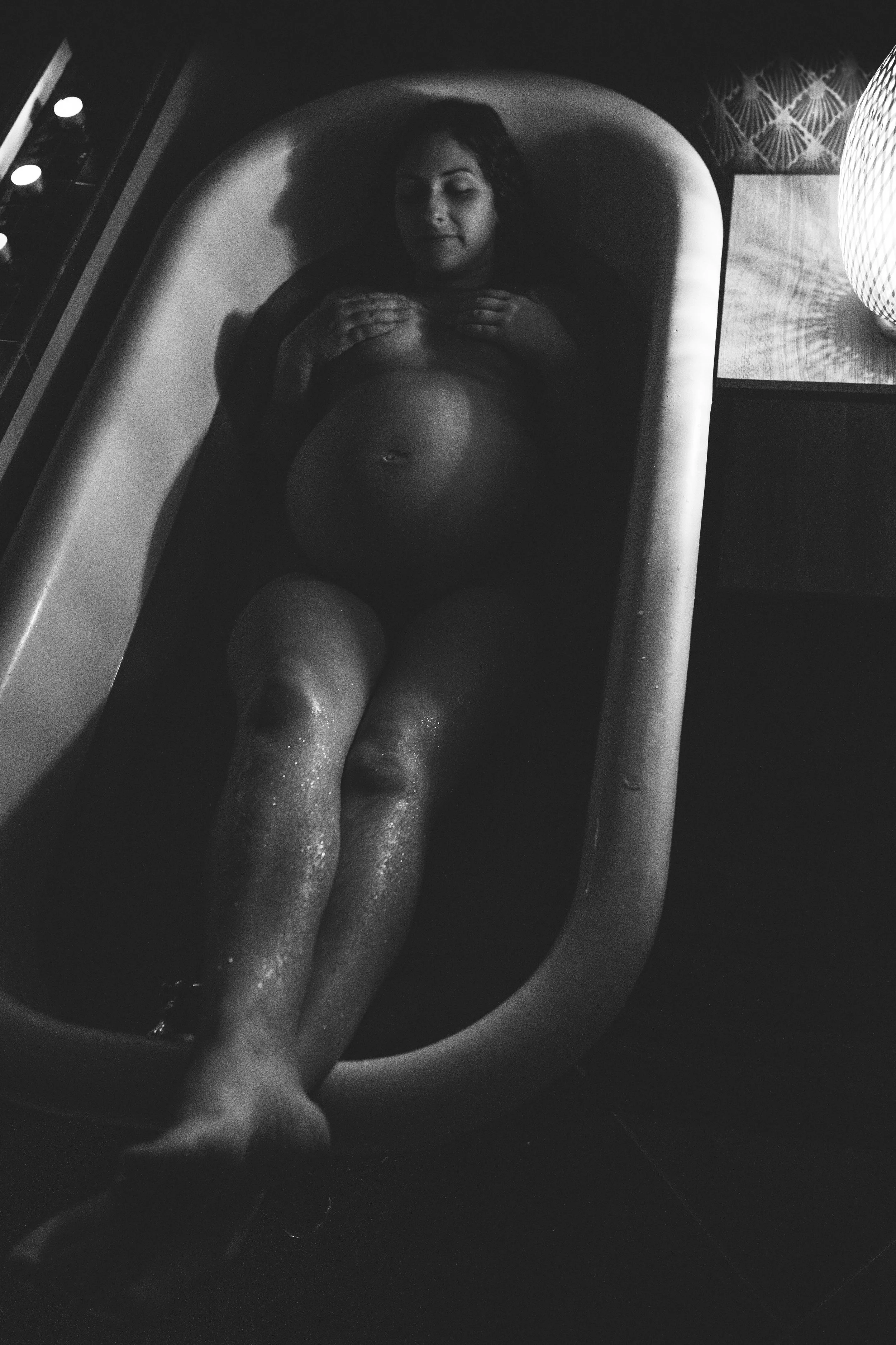 A woman lying in a bathtub with her hands on her chest, with water on her legs, and lightly closed eyes, in a black-and-white photo with low lighting.