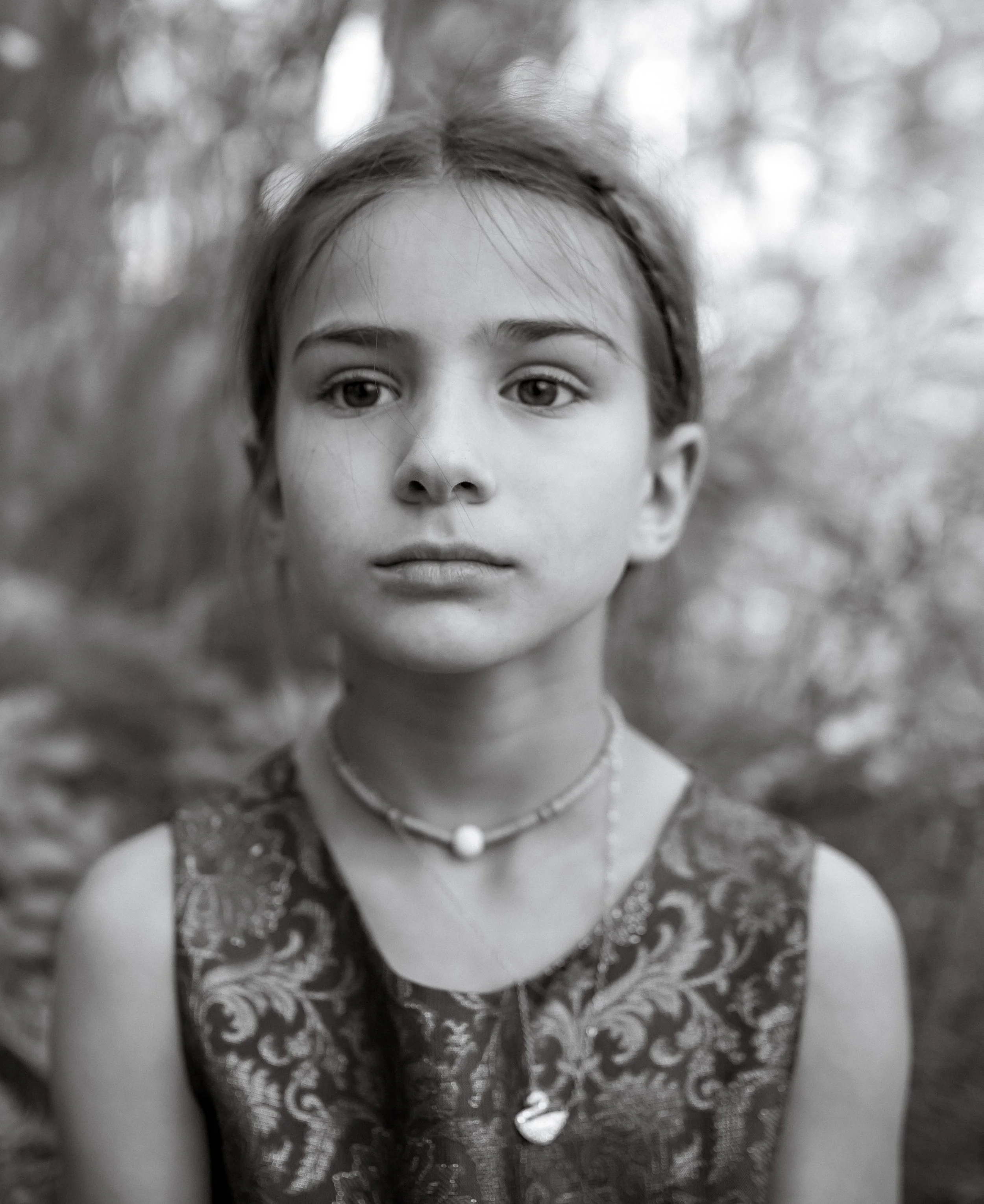 Black and white photo of a young girl in traditional attire, wearing a lace dress and a necklace, with outdoor blurred background.
