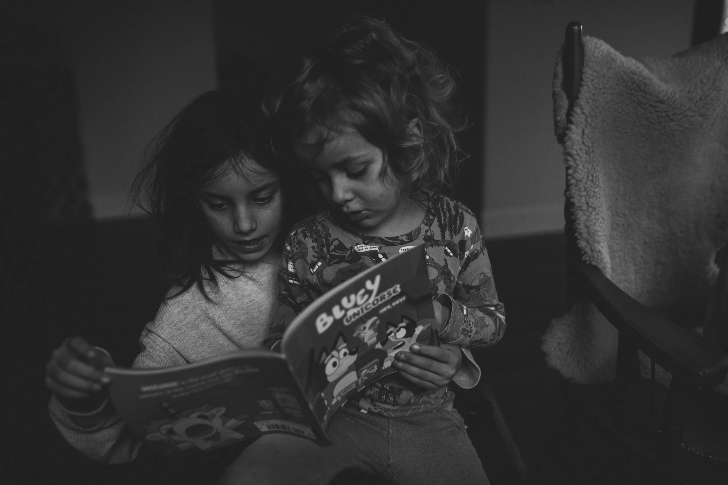 Two young children, a girl and a boy, sitting close together and reading a comic book in black and white. The girl has curly hair and is leaning toward the boy, who is wearing patterned pajamas with cartoon characters.