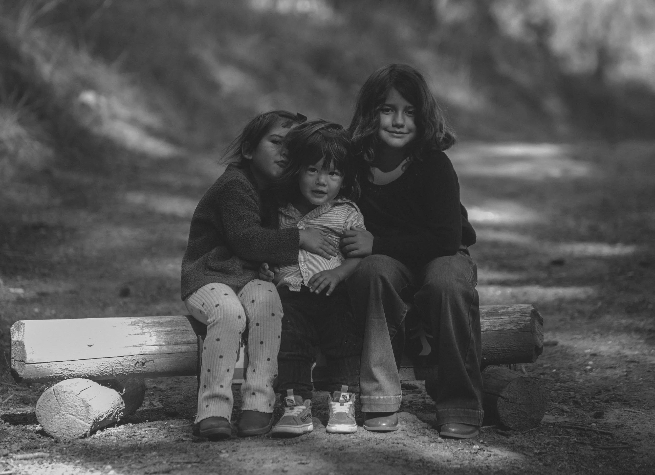 Three children sitting on a bench outdoors in a black and white photo. The children are close together, with the girl on the right smiling, the girl on the left leaning in, and the boy in the middle looking at the camera. There is a soccer ball in fr