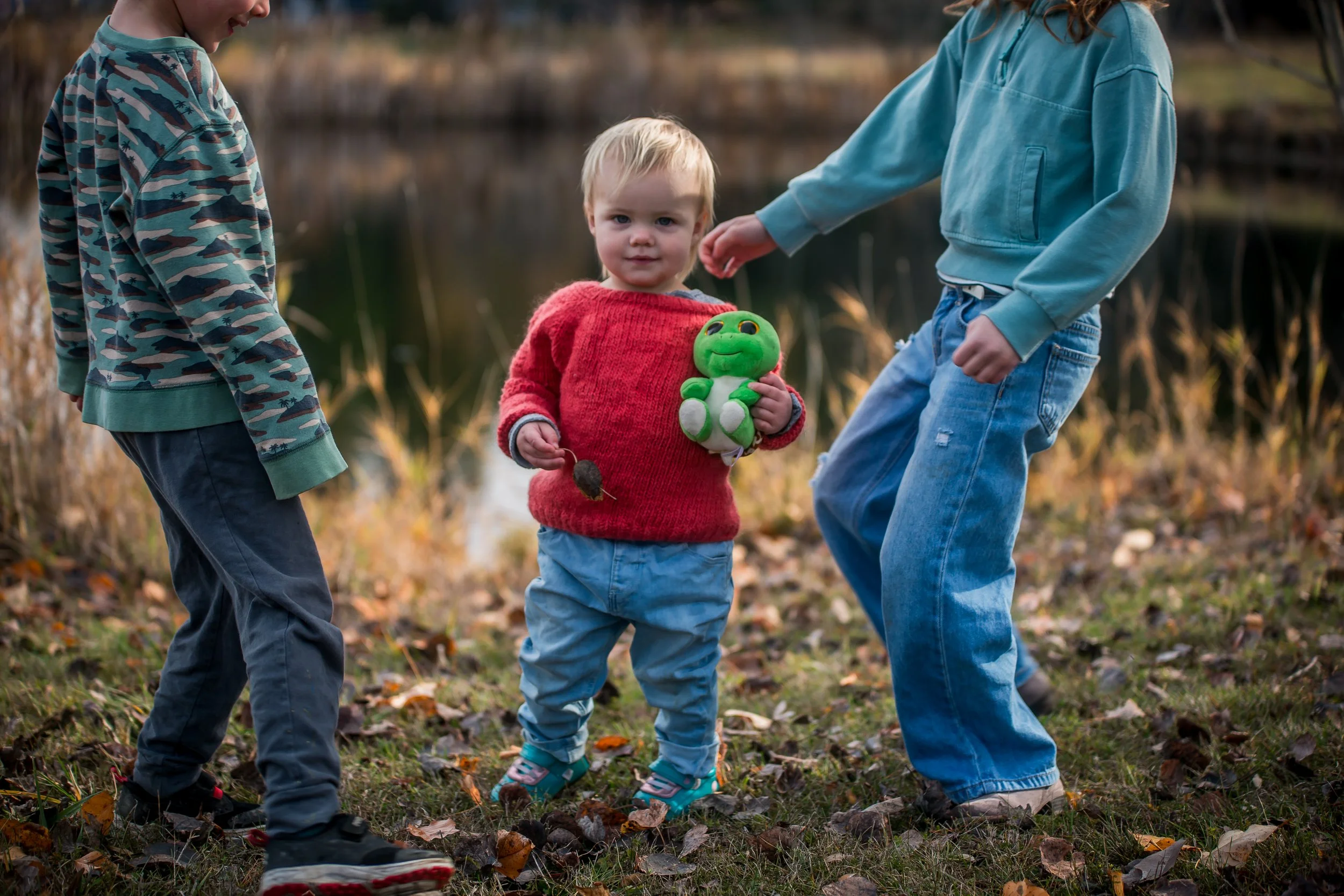 Three children playing outdoors near a lake in fall, holding a green stuffed animal, with one child in a red sweater and two in blue and camouflage hoodies.