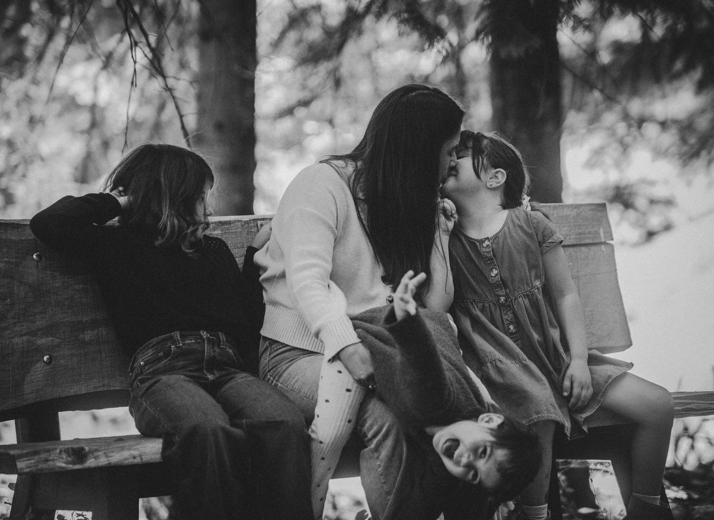 A woman and two children sitting on a park bench, sharing a kiss. One girl is sitting next to them, resting her head on her hand. The scene is outdoors with trees in the background.