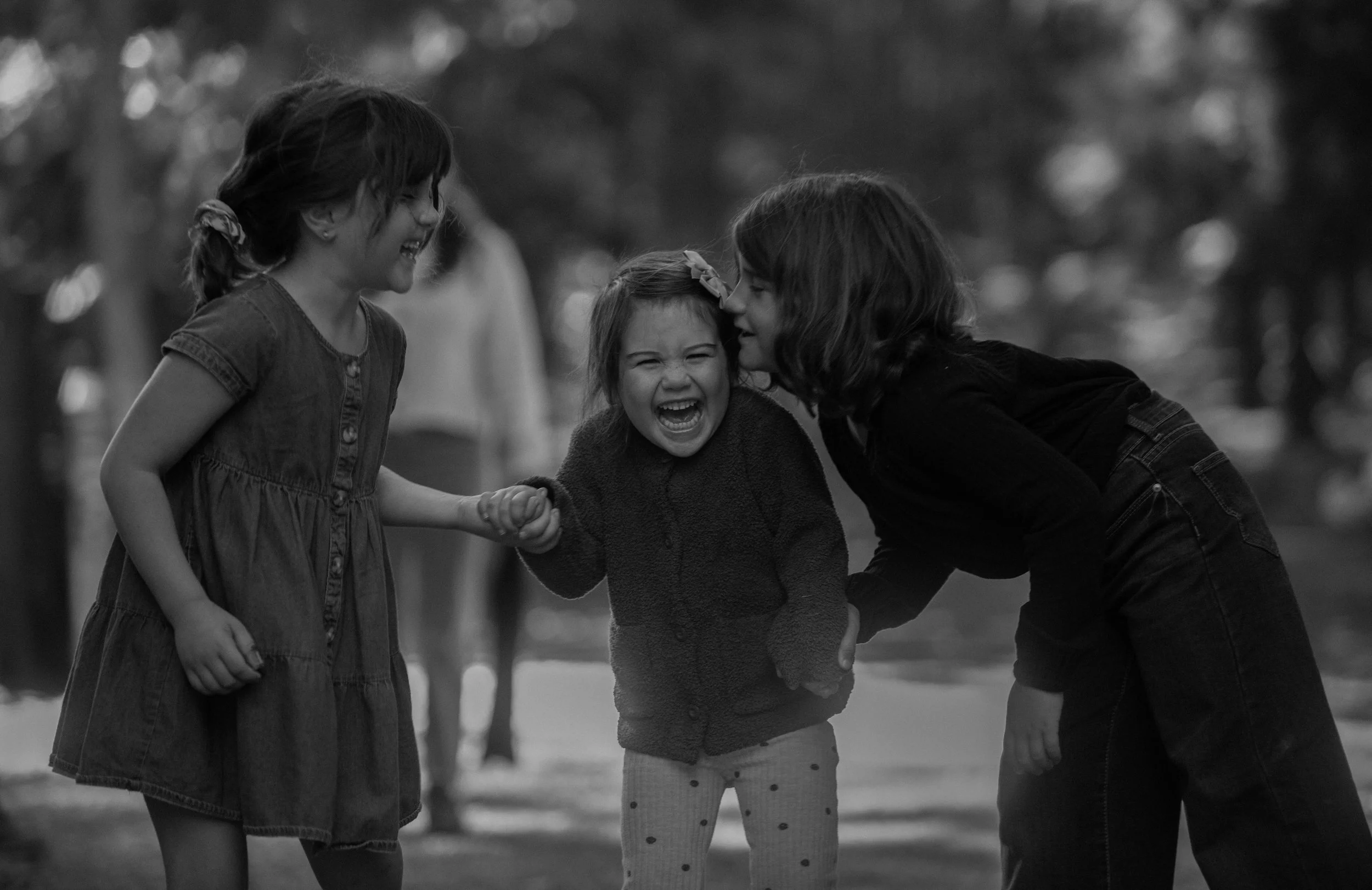 Three young girls laughing and playing together outside, holding hands, in a park with blurred trees in the background.