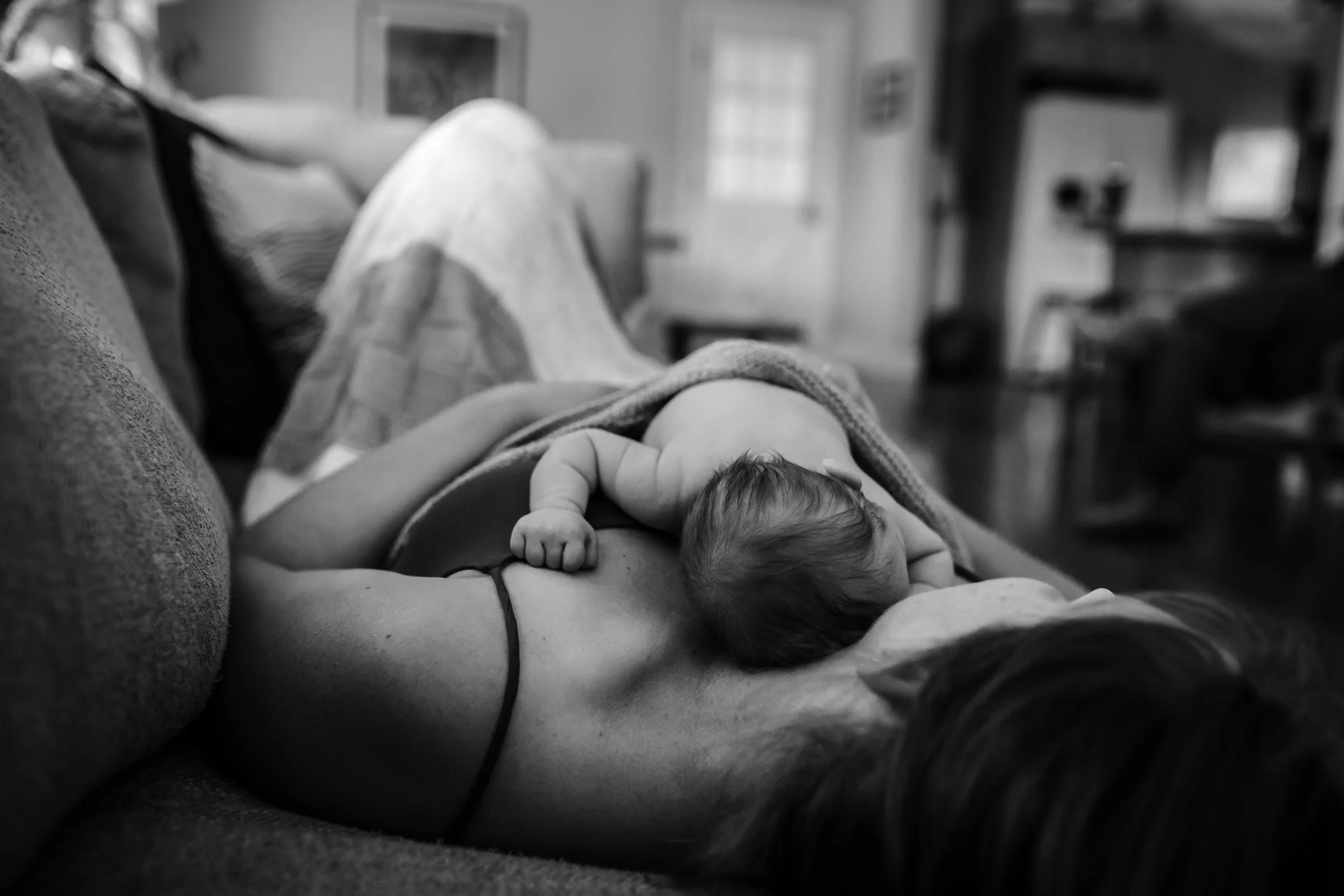 A person lying on a couch with a baby close to their chest, inside a living room with shelves and a window.