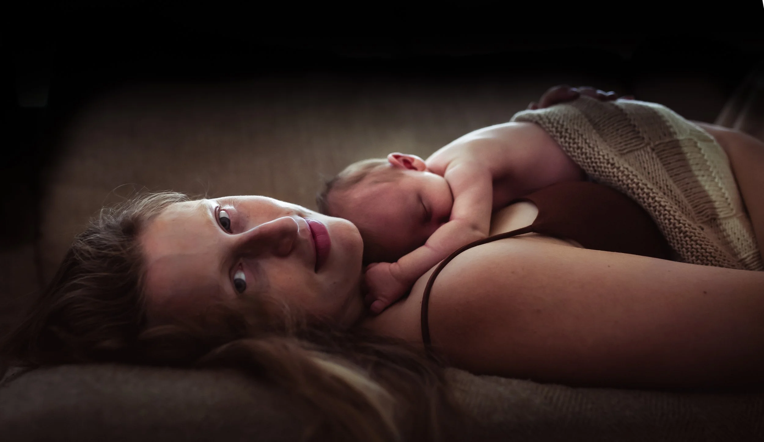 A woman lying on her back with a baby sleeping on her chest. The woman has red hair and is looking up, while the baby is wrapped in a beige and brown knitted blanket.
