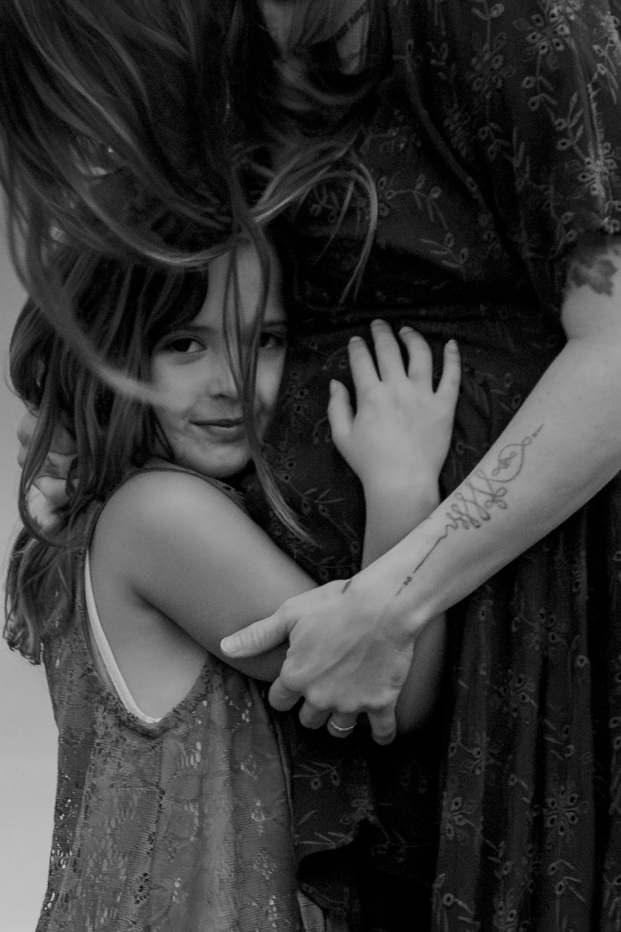 A woman with long hair embraces a man. The woman's face is visible, showing a gentle smile, and she is looking into the camera. The woman's arm and hand rest on the man's back. The woman is wearing a lace dress with a cut-out shoulder design. The man, whose face is not shown, has a tattoo on his forearm. The image is in black and white.