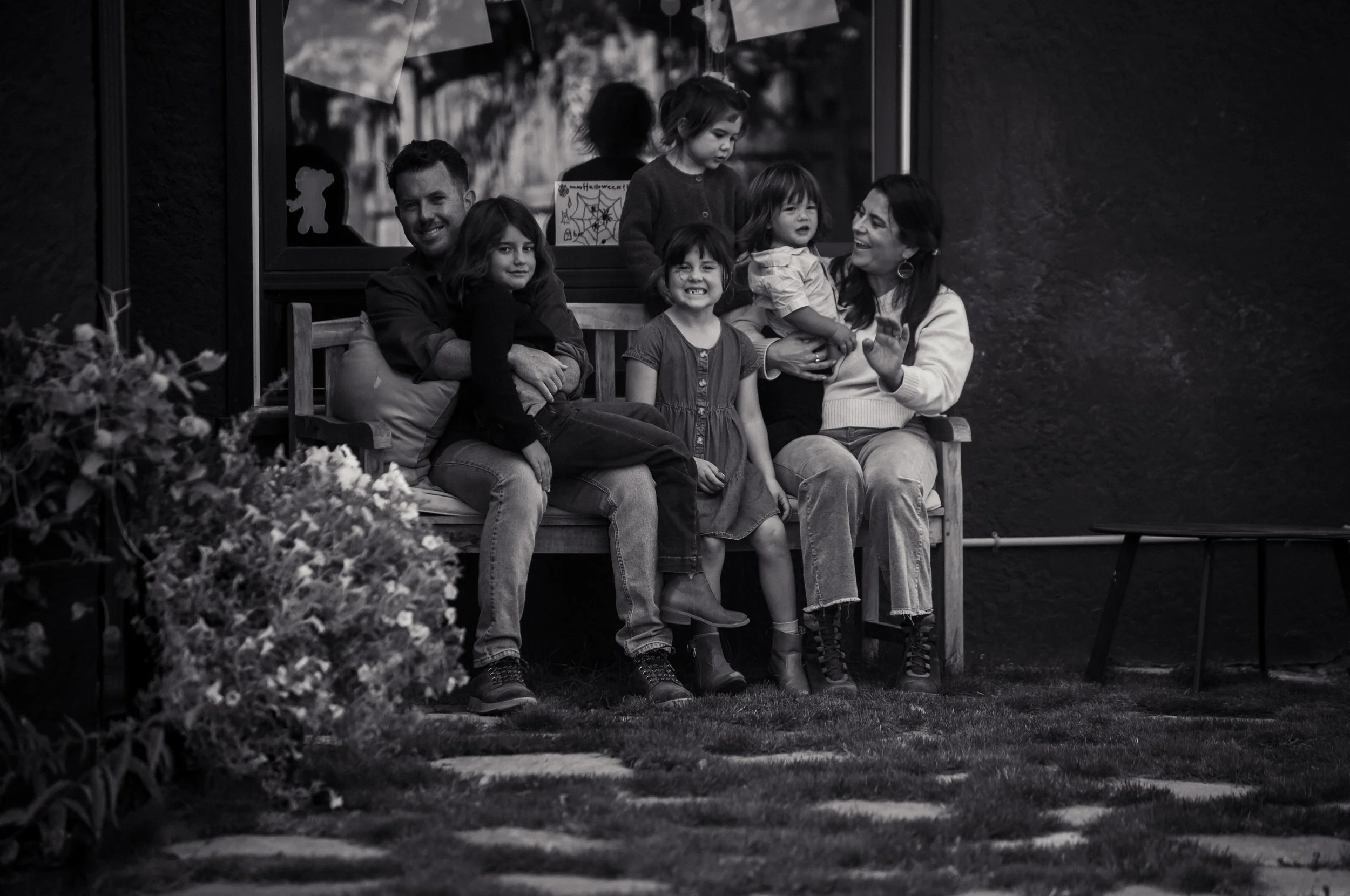 A group of children and a woman sitting on a wooden bench outside near a window decorated with Halloween drawings, smiling and talking.