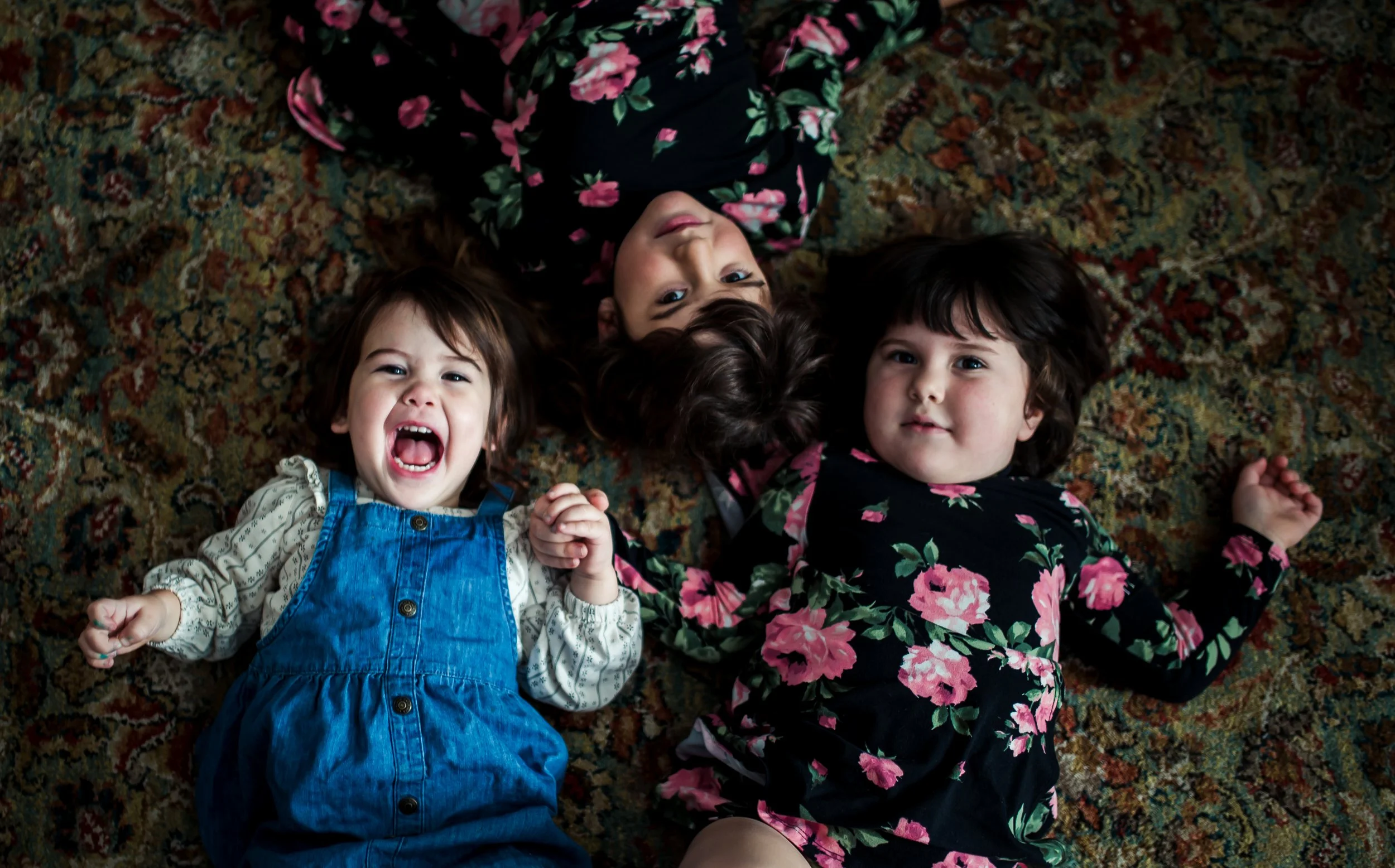 Three young girls lying on a patterned carpet, smiling and looking up at the camera. The girl on the left has brown hair and is wearing a white shirt with blue overalls, the girl in the middle has dark hair and is wearing a black floral dress, and th