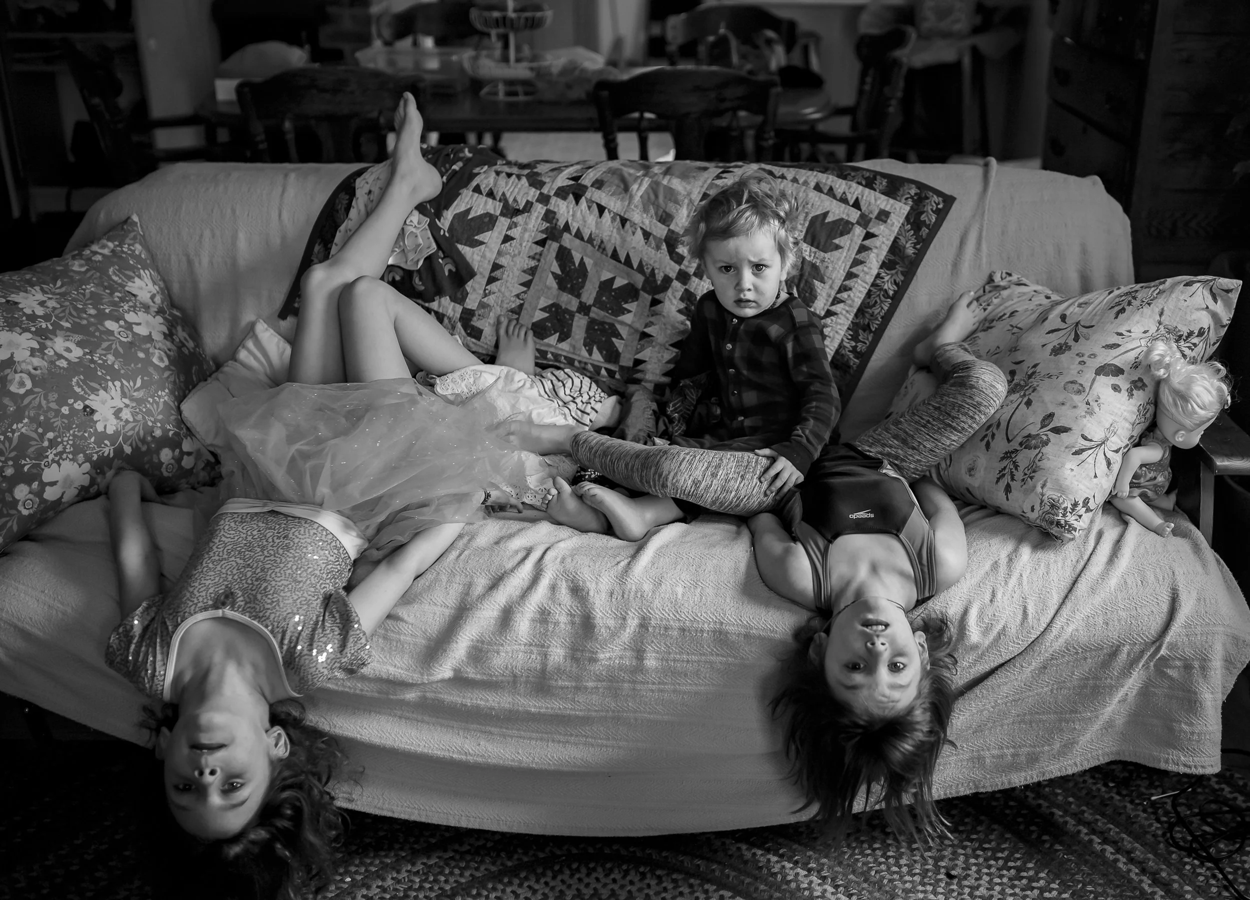 A black-and-white photo of four children on a sofa, two with their heads upside down, one lying on her back with feet up, and a young boy sitting up, looking at the camera with a serious expression. There are pillows and a blanket on the sofa, and a 