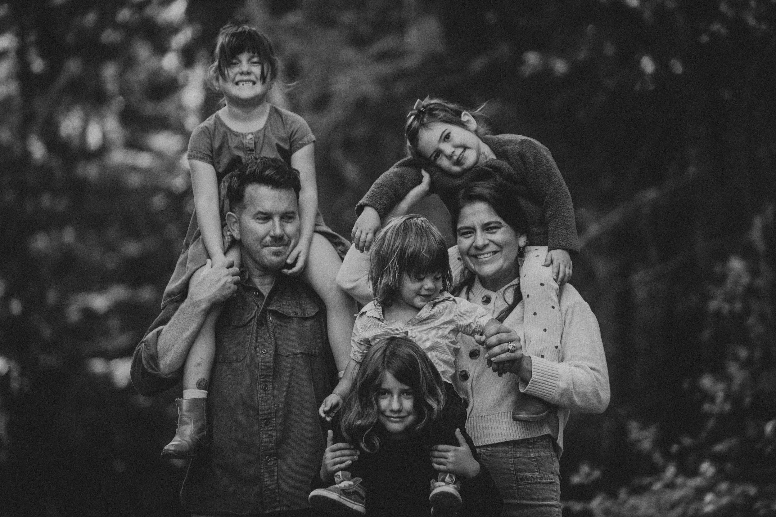 A black and white photo of a happy family outdoors, with children sitting on parents' shoulders, smiling and enjoying a moment together.