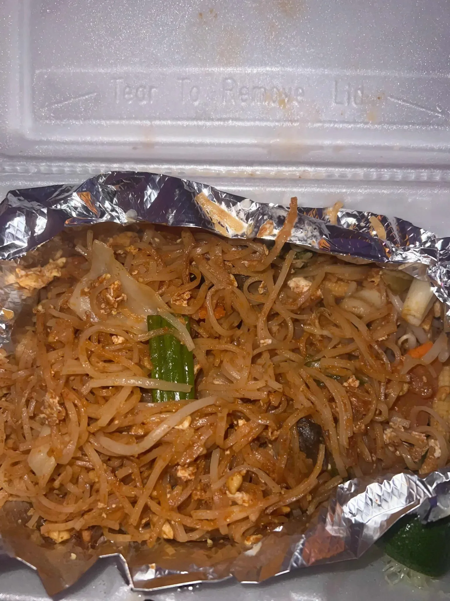 A takeout container filled with stir-fried noodles, vegetables, and scrambled eggs, with a small packet of soy sauce beside it.
