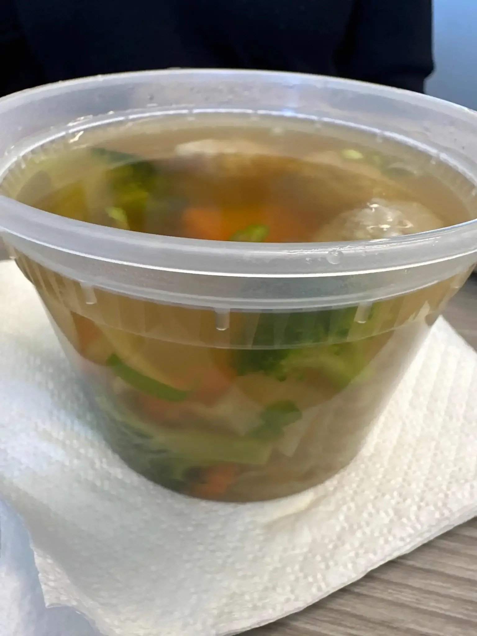 A clear plastic container filled with soup or broth, containing vegetables and possibly meat, placed on a white textured surface.