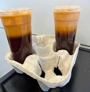 Two plastic cups of iced coffee with dark coffee at the bottom and a layer of lighter cream or milk on top, sitting in a gray cardboard drink carriers.