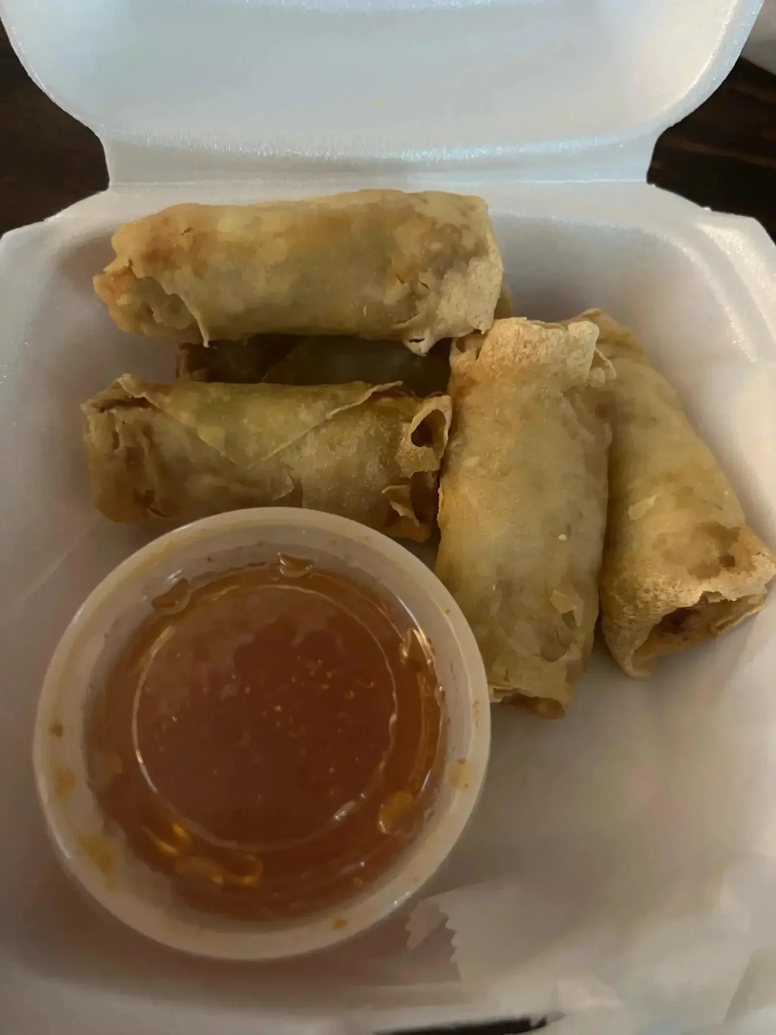 A white foam container holding several fried spring rolls with a small cup of dipping sauce.