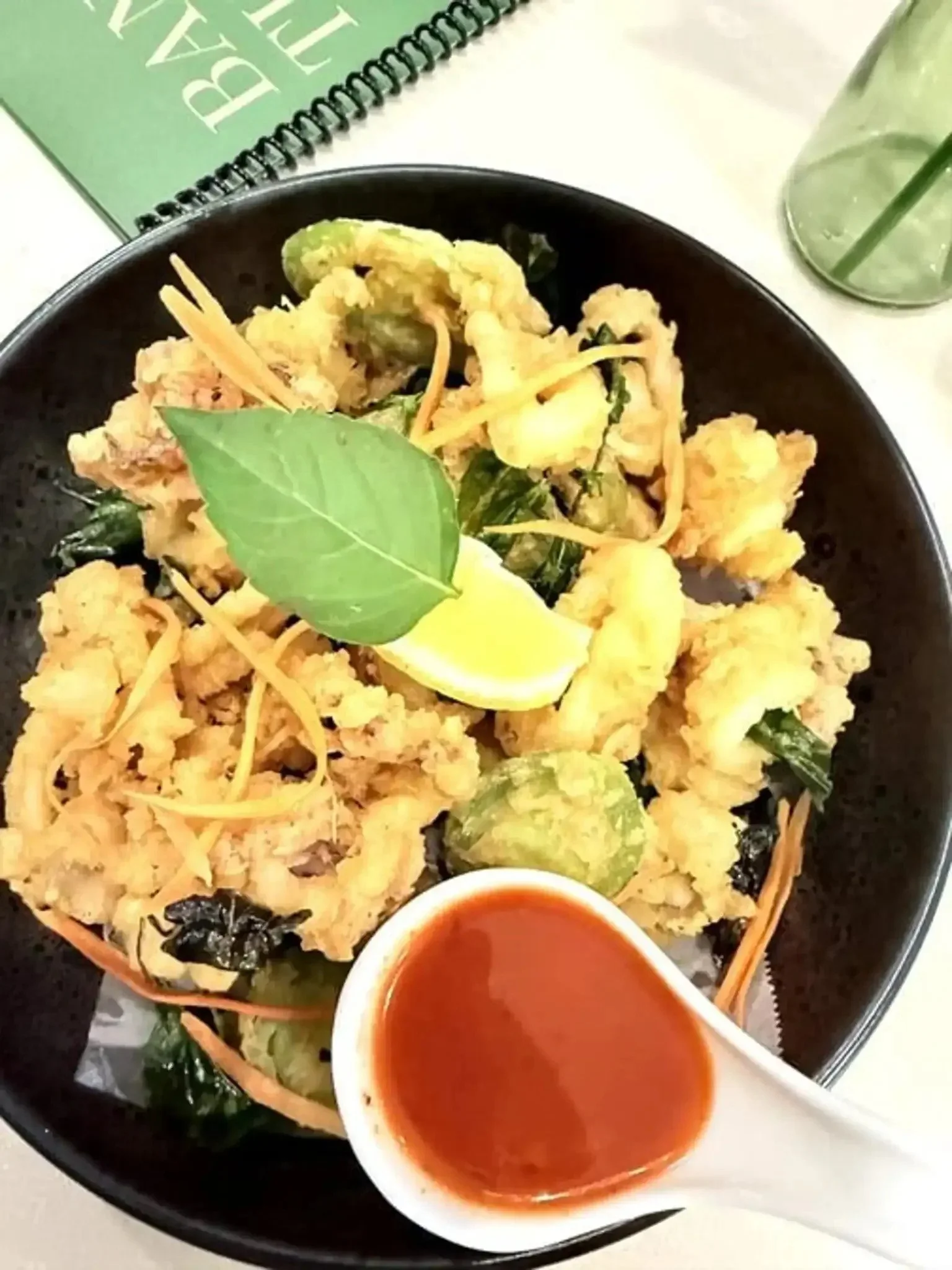 Close-up of a black bowl with assorted tempura vegetables, a lemon wedge, fresh herbs, and a small cup of dipping sauce, alongside a tall glass of iced water with a straw and a green spiral notebook.