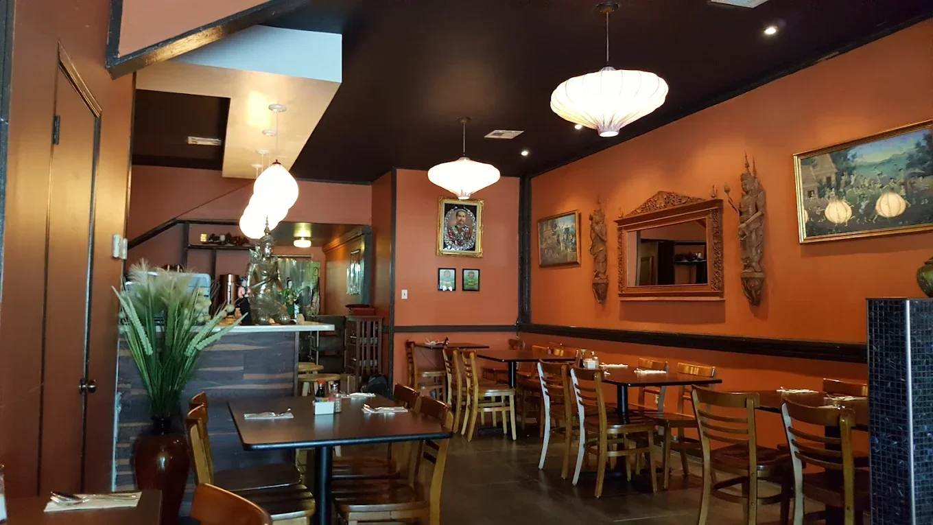 Interior of a restaurant with wooden chairs and tables, framed artwork and sculptures on a warm-colored wall, covered pendant lights, and a plant in a vase.