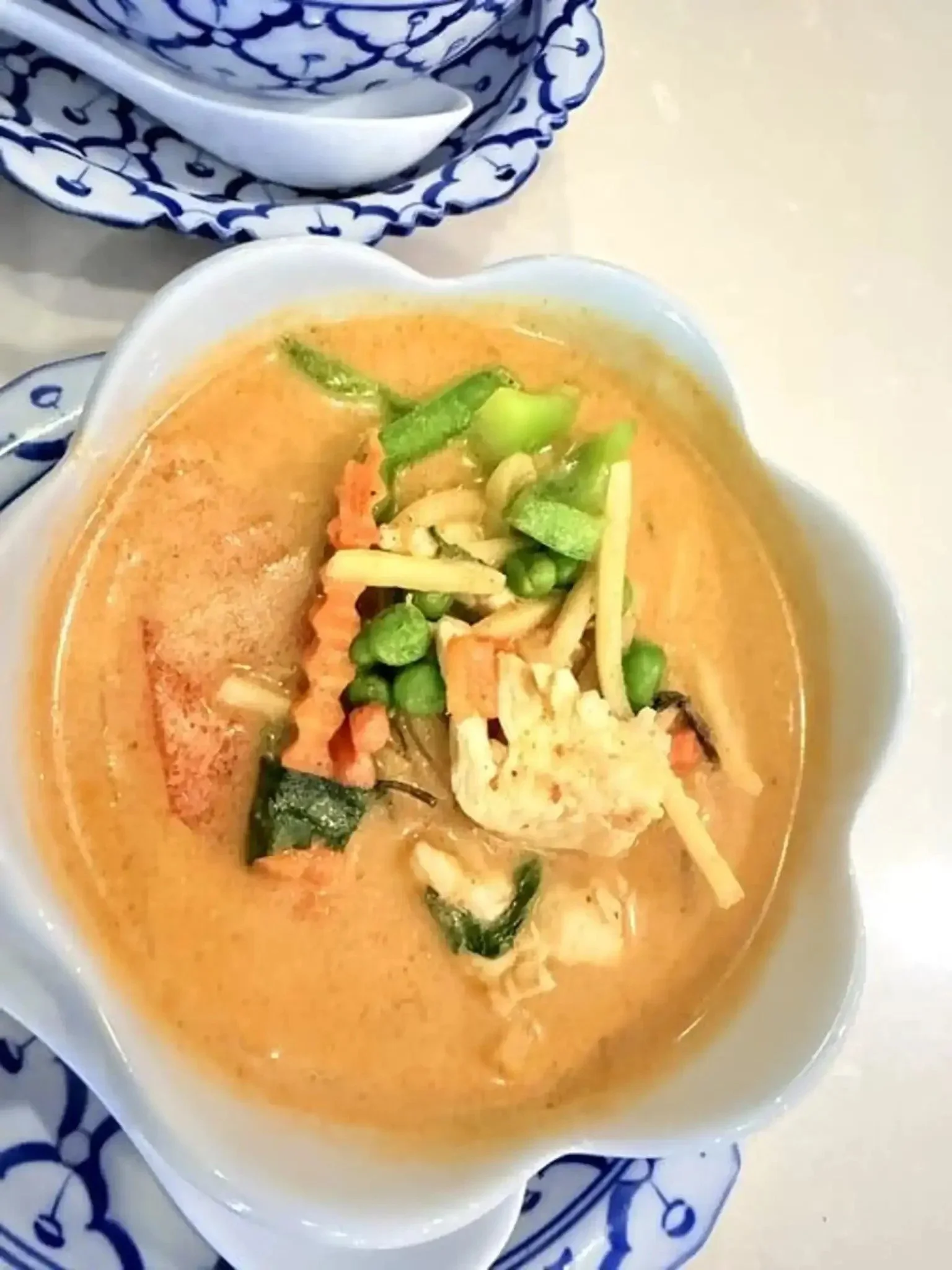 A bowl of creamy, orange-colored soup garnished with chopped green herbs and vegetables, placed on a decorative blue and white plate with a matching spoon and napkin.