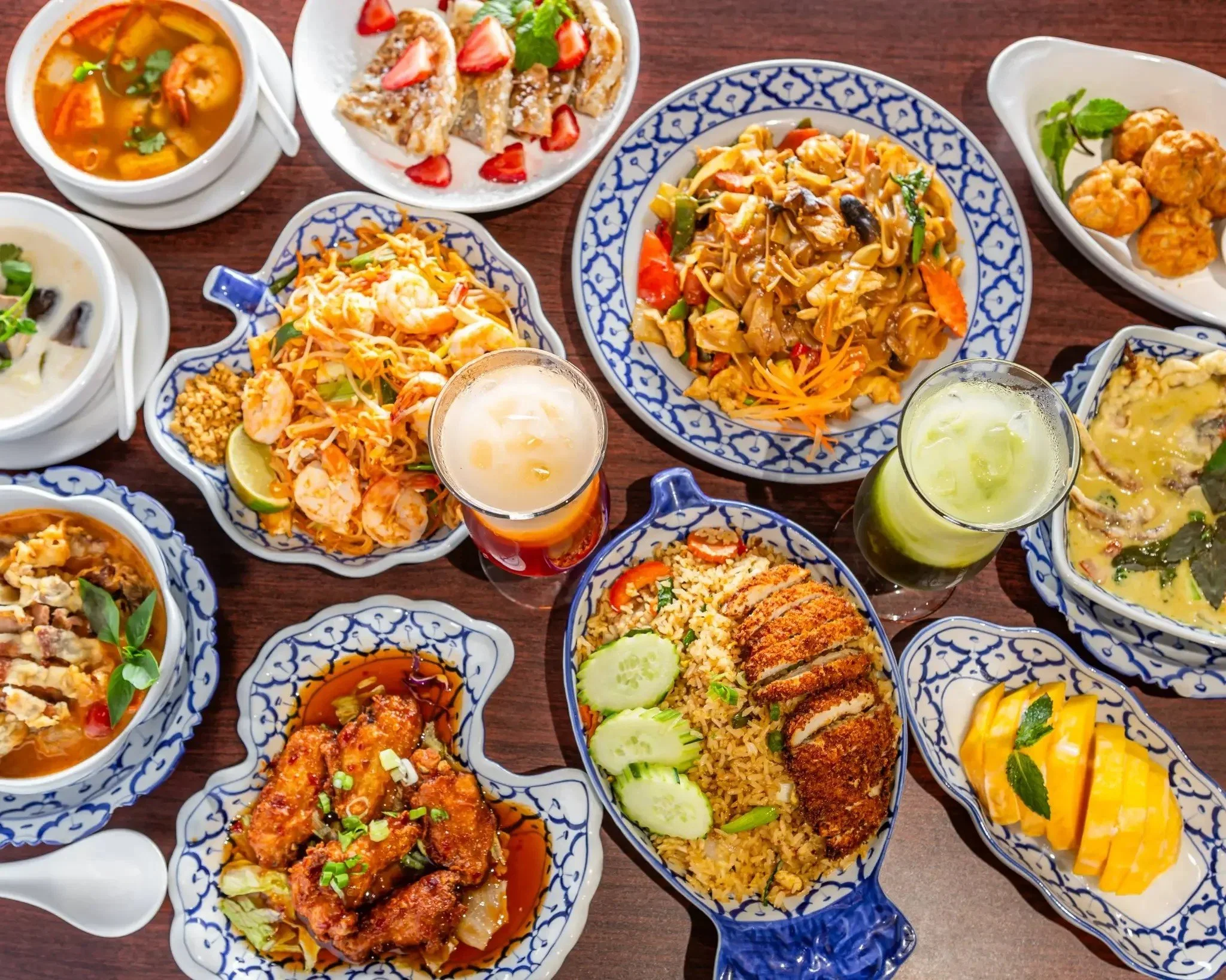 Assorted Thai dishes served in blue and white patterned bowls, including noodle stir fry, fried chicken, shrimp salad, and vegetable soups, with side dishes and drinks on a wooden table.