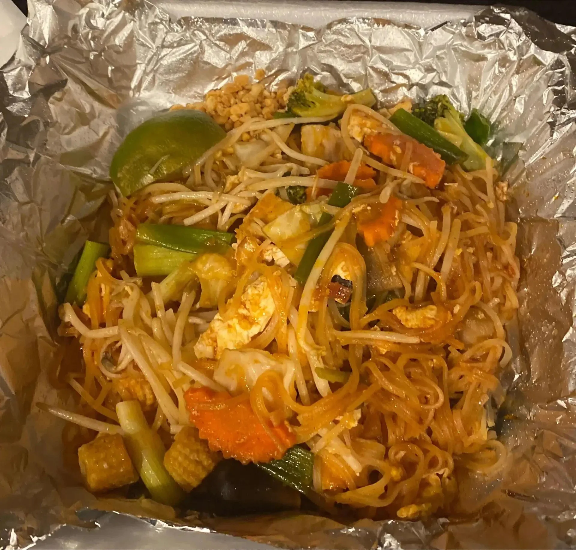 Close-up of cooked noodles with vegetables, lime wedge, and chopped peanuts on aluminum foil.