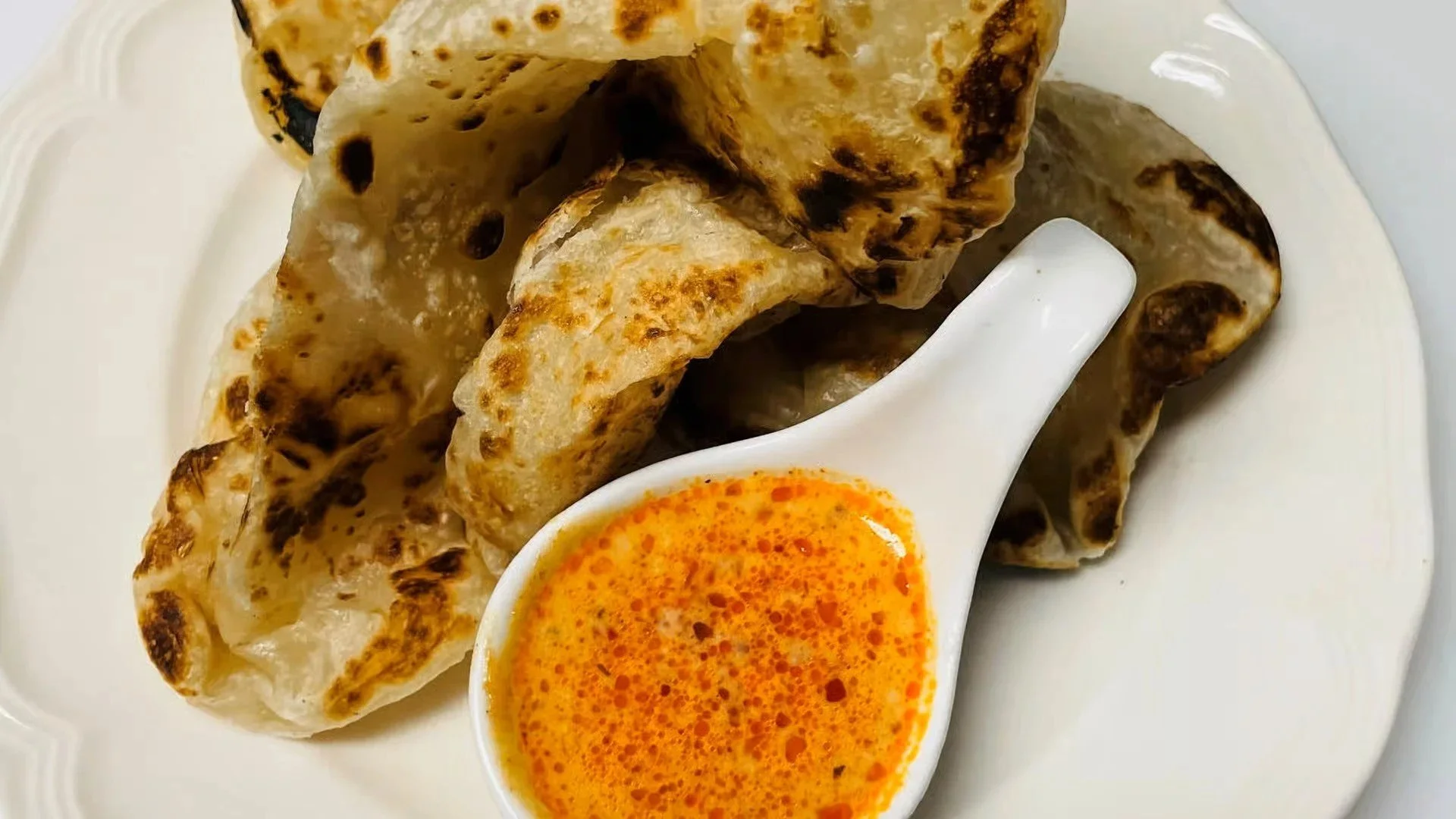 Close-up of grilled flatbread pieces served with a small bowl of orange-colored dipping sauce on a white plate.