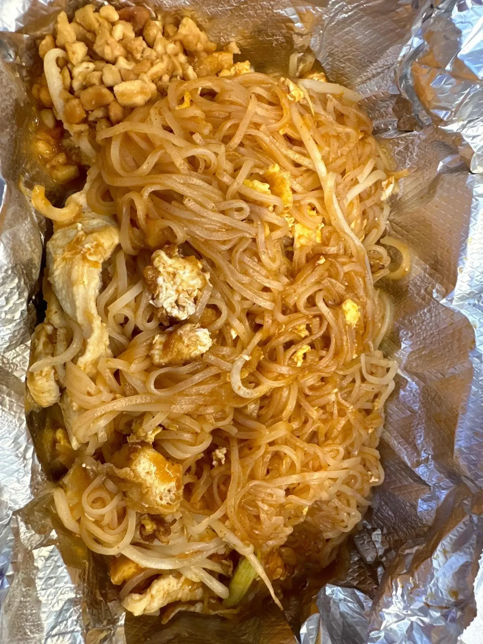 Close-up of fried noodles topped with scrambled eggs, bean sprouts, and pieces of fried chicken, all on aluminum foil.
