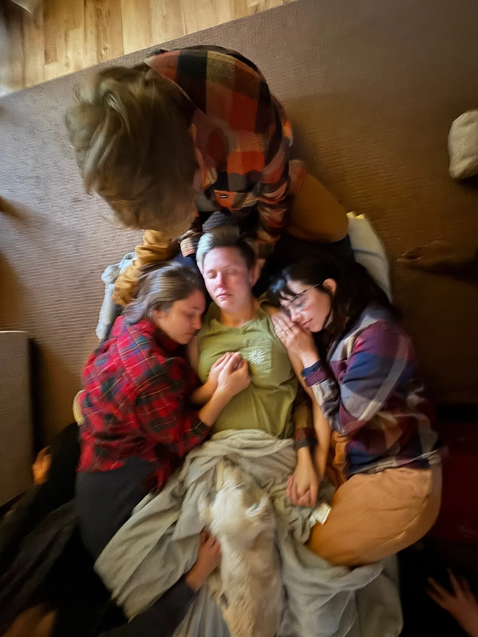 A group of four people are gathered closely together on a carpeted floor, sleeping and resting. They are lying in a circle, with some resting their heads on each other or holding hands, appearing peaceful and comfortable.