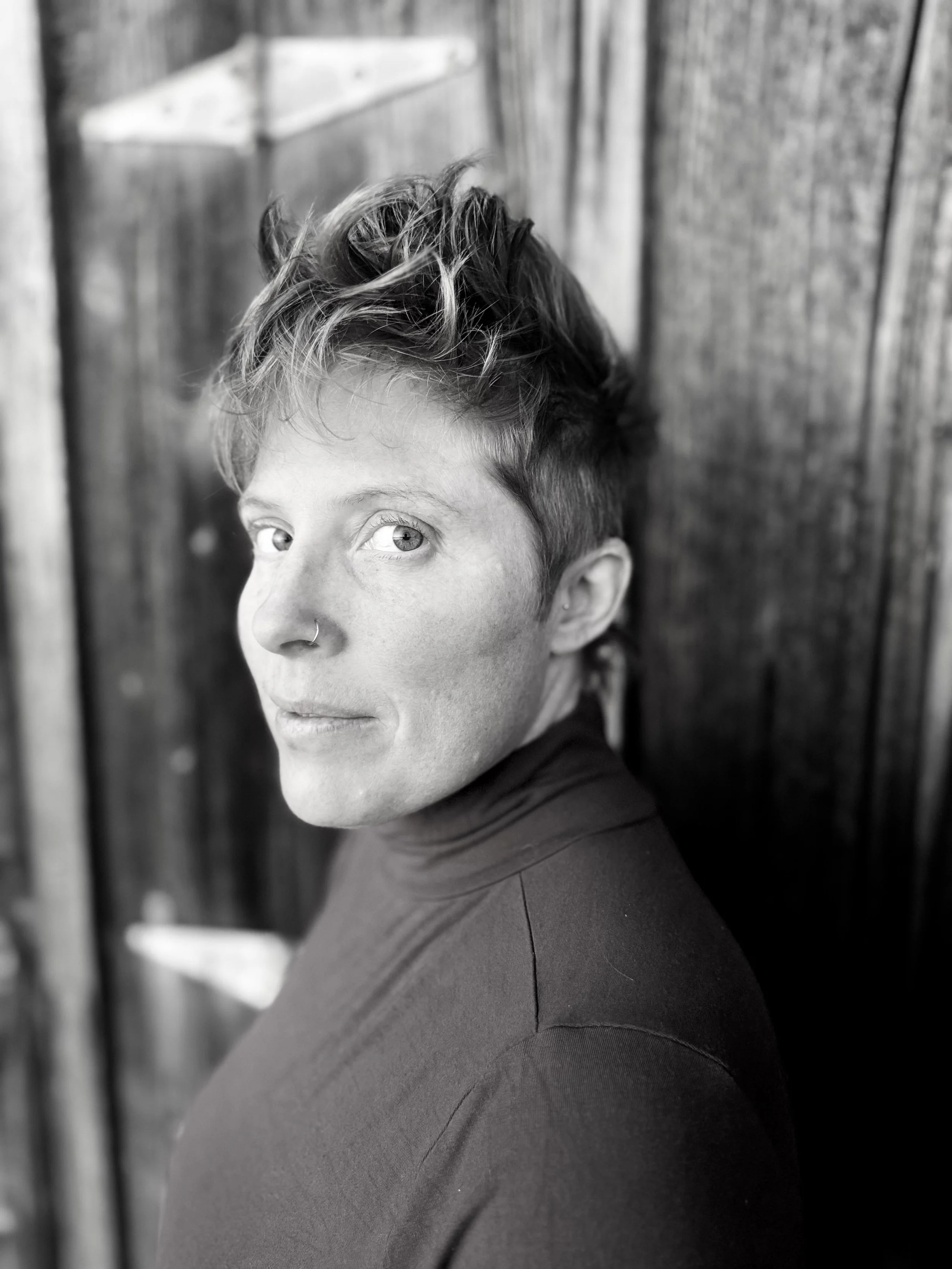 Black and white photo of a person with short hair and a nose piercing, wearing a dark top, standing in front of a wooden wall or fence.