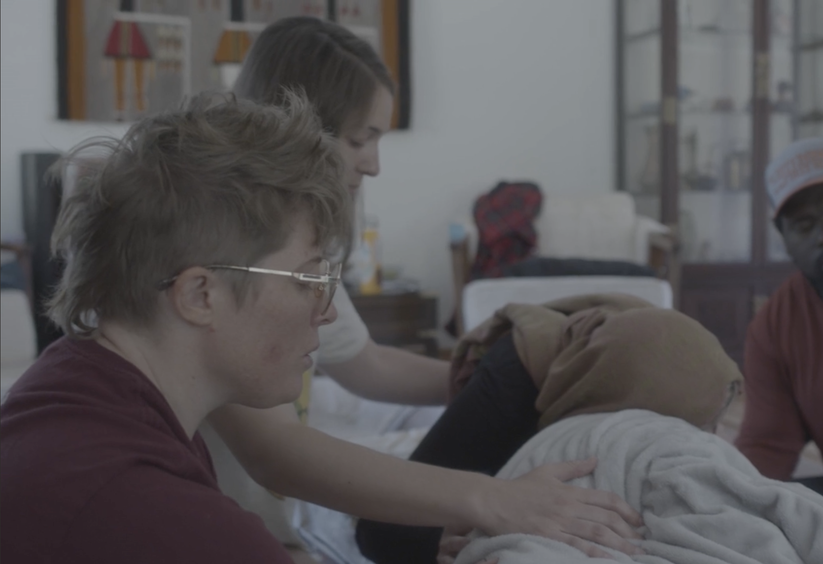 Group of people in a caregiving setting, with one individual lying on a bed receiving assistance from others, including a woman tending to the person and others observing, in a room with shelves and decorative items.