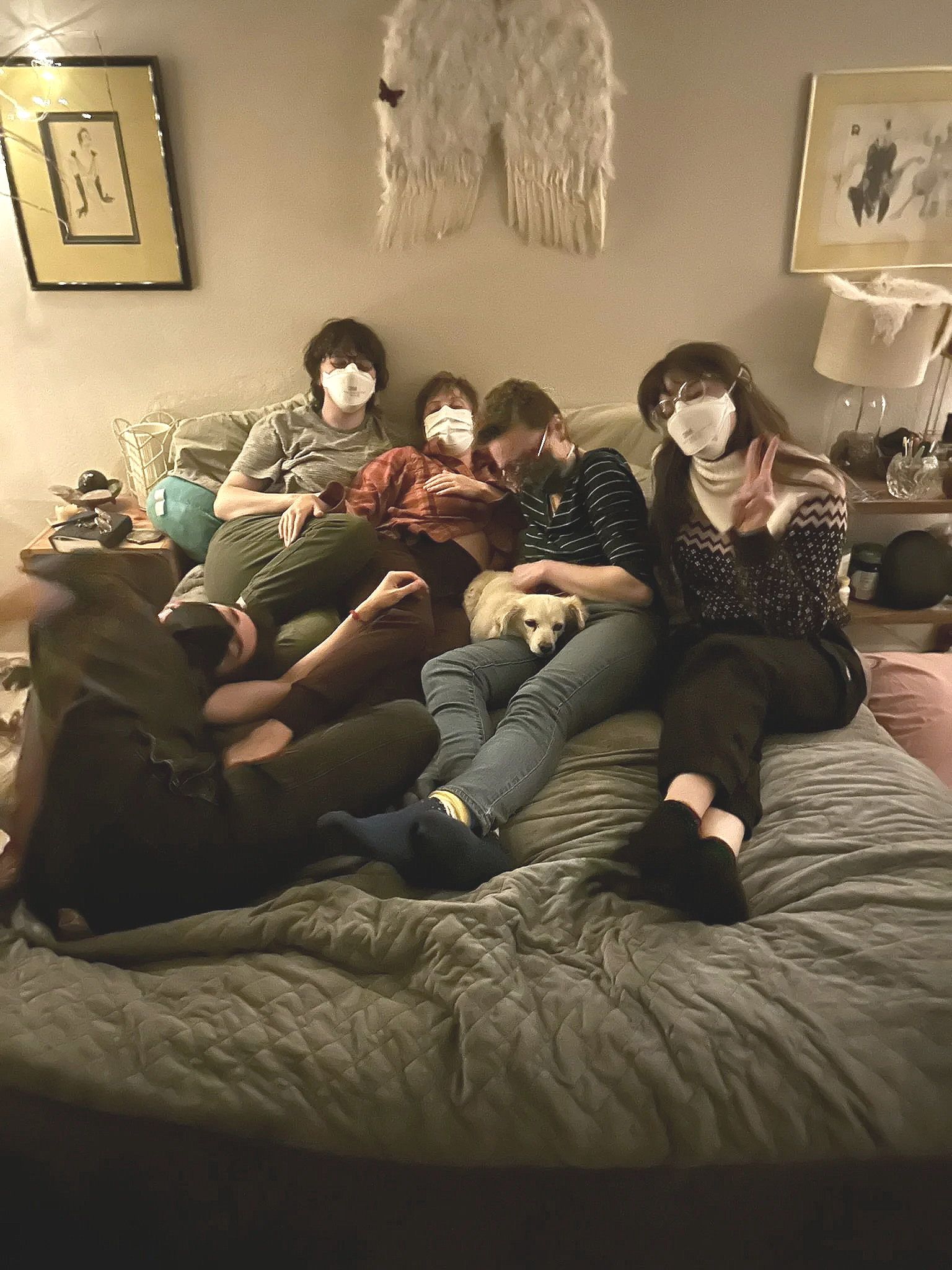 Four people, all wearing masks, are sitting on a bed in a cozy room. They are posing for a photo, some making peace signs. There are framed pictures and a lamp on the nightstand, and a dog is lying on the bed.