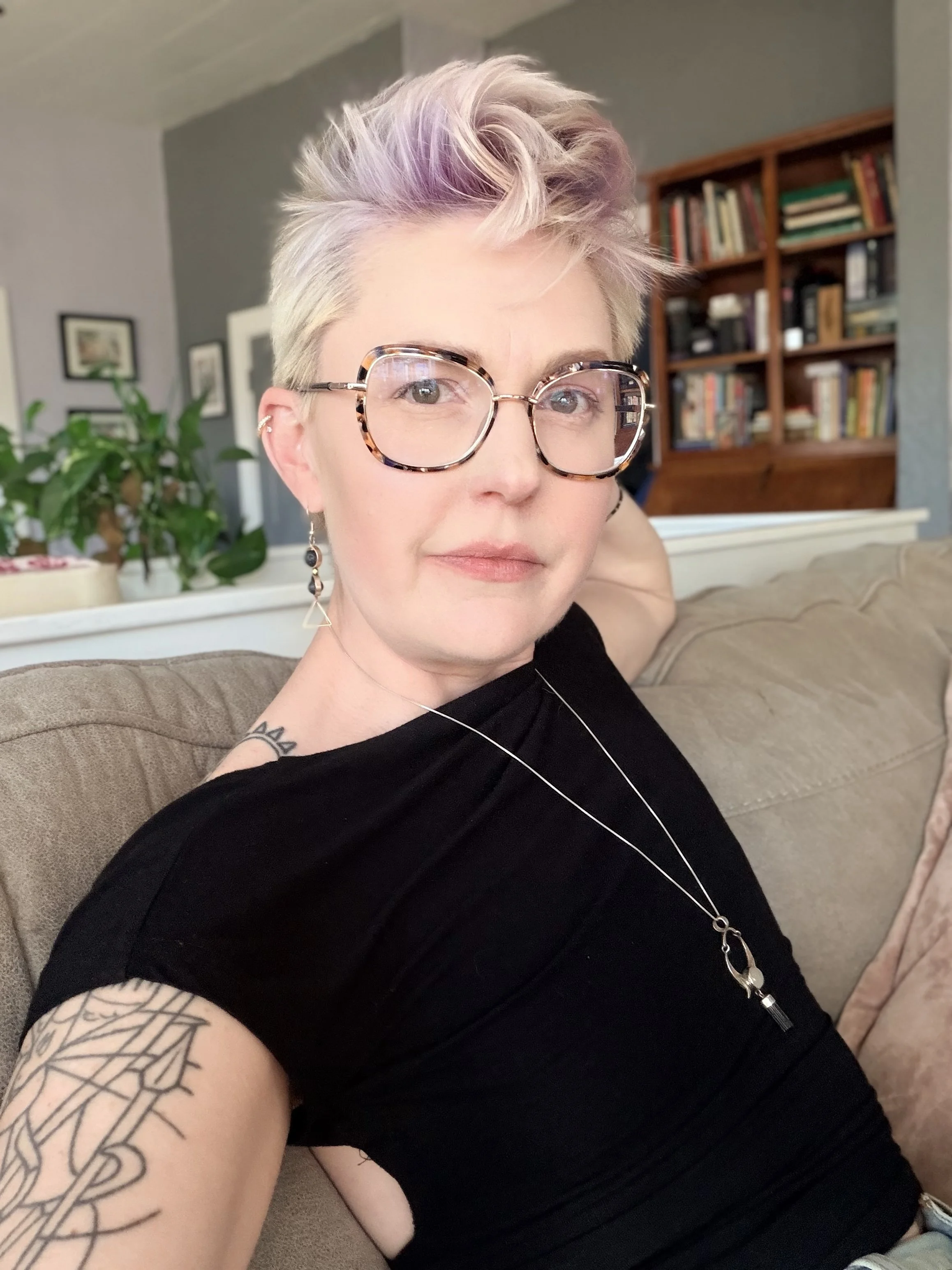 A woman with short, platinum blonde hair with light pink highlights, large tortoiseshell glasses, a black shirt, and geometric earrings, sitting on a beige couch in a living room with a wooden bookshelf and potted plant in the background.