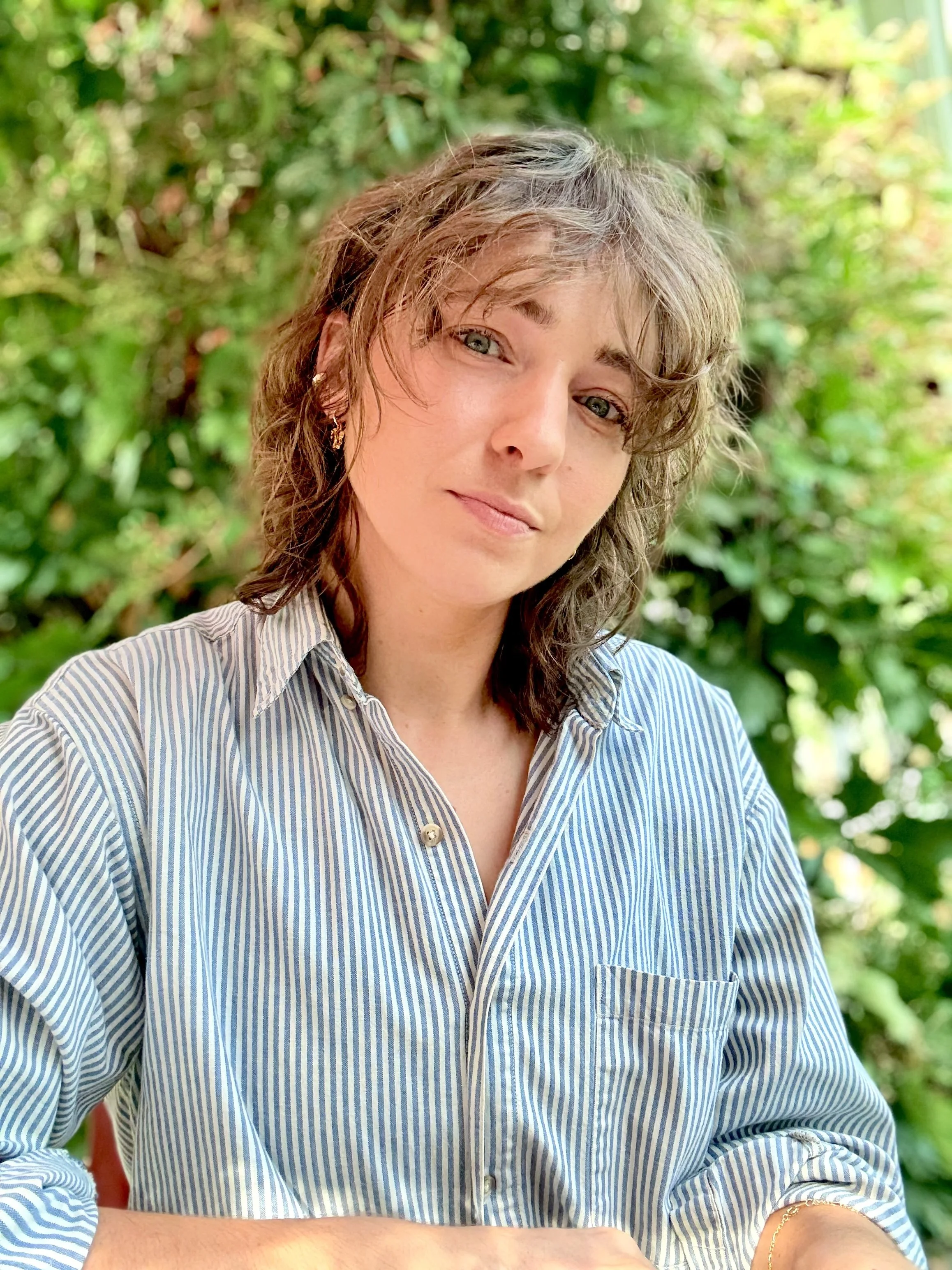 Close-up of a woman with wavy brown hair and blue eyes, wearing a light blue and white striped shirt, sitting outdoors with green foliage in the background.