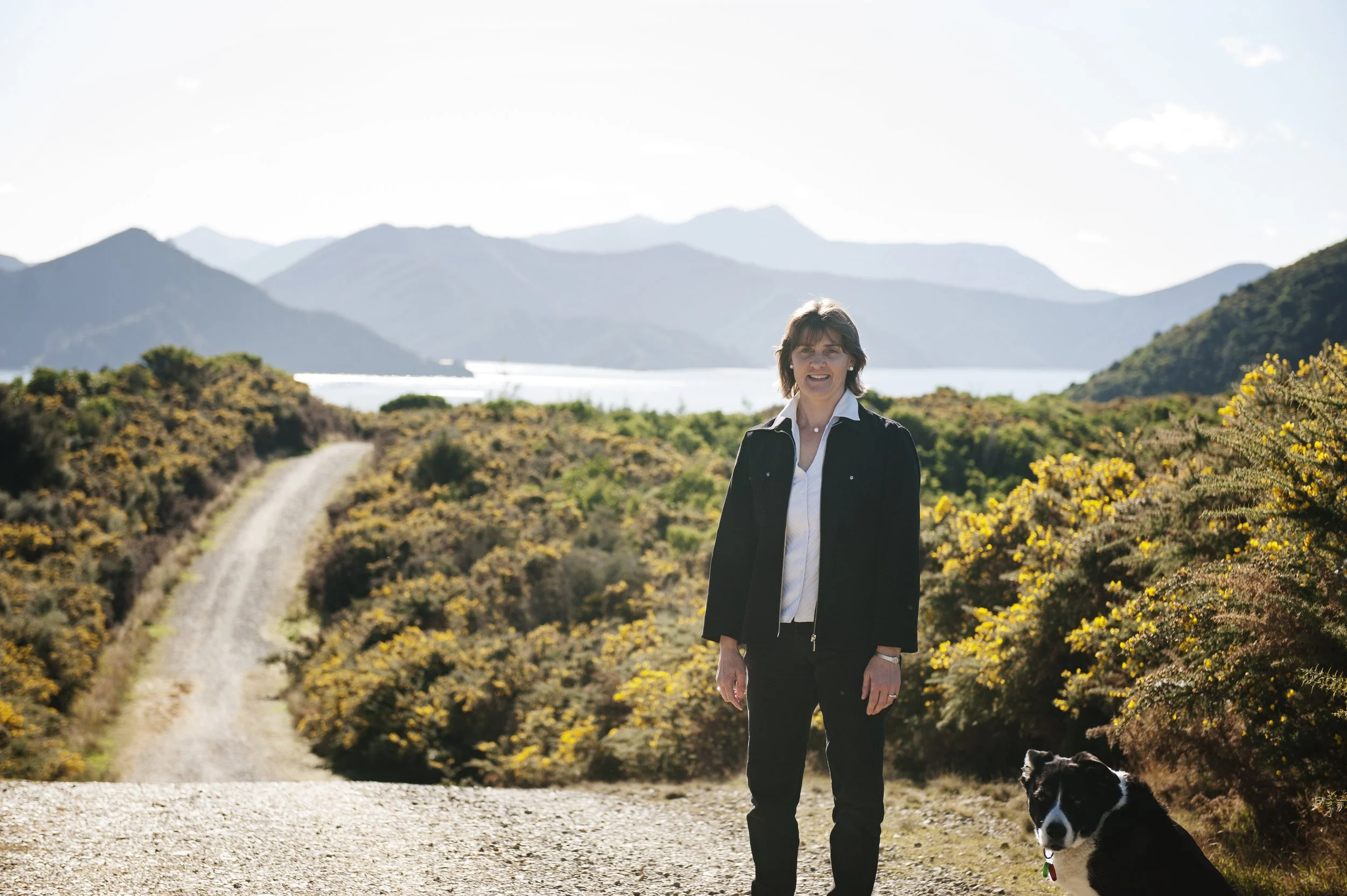 A woman standing on a dirt path in a mountainous landscape with yellow flowering bushes, a lake, and mountains in the background, accompanied by a black and white dog.